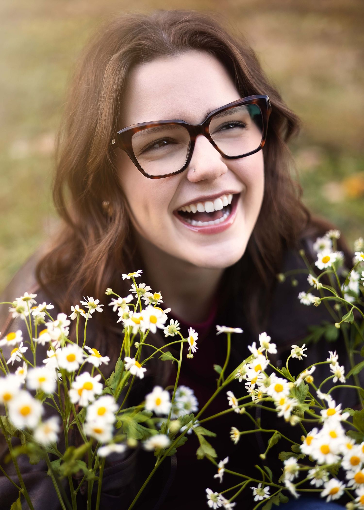 Smiling young woman with glasses holding white daisy flowers outdoors in a bright, cheerful portrait photo.