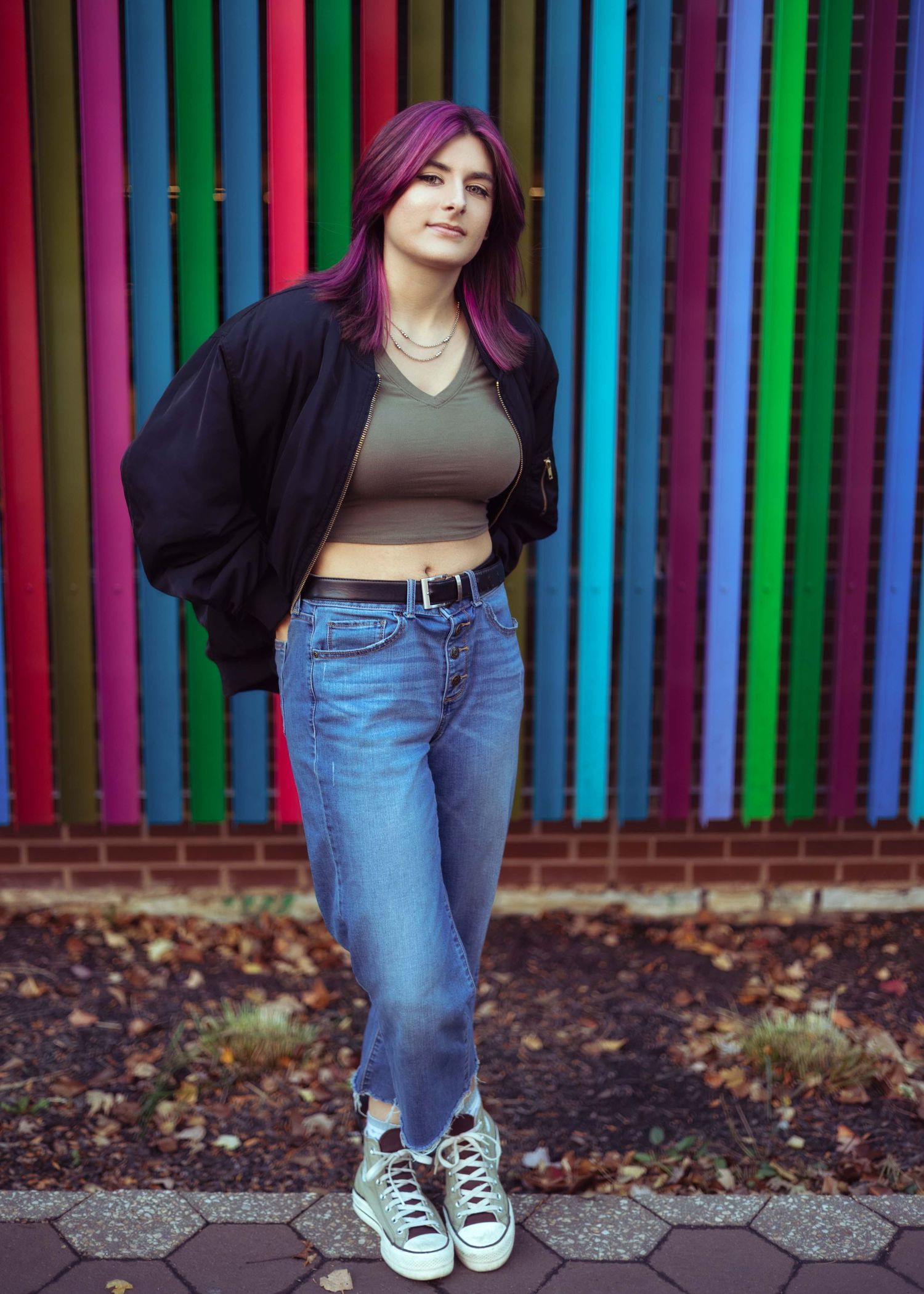 Young woman with purple hair in black bomber jacket, crop top, and jeans poses before colorful striped wall.