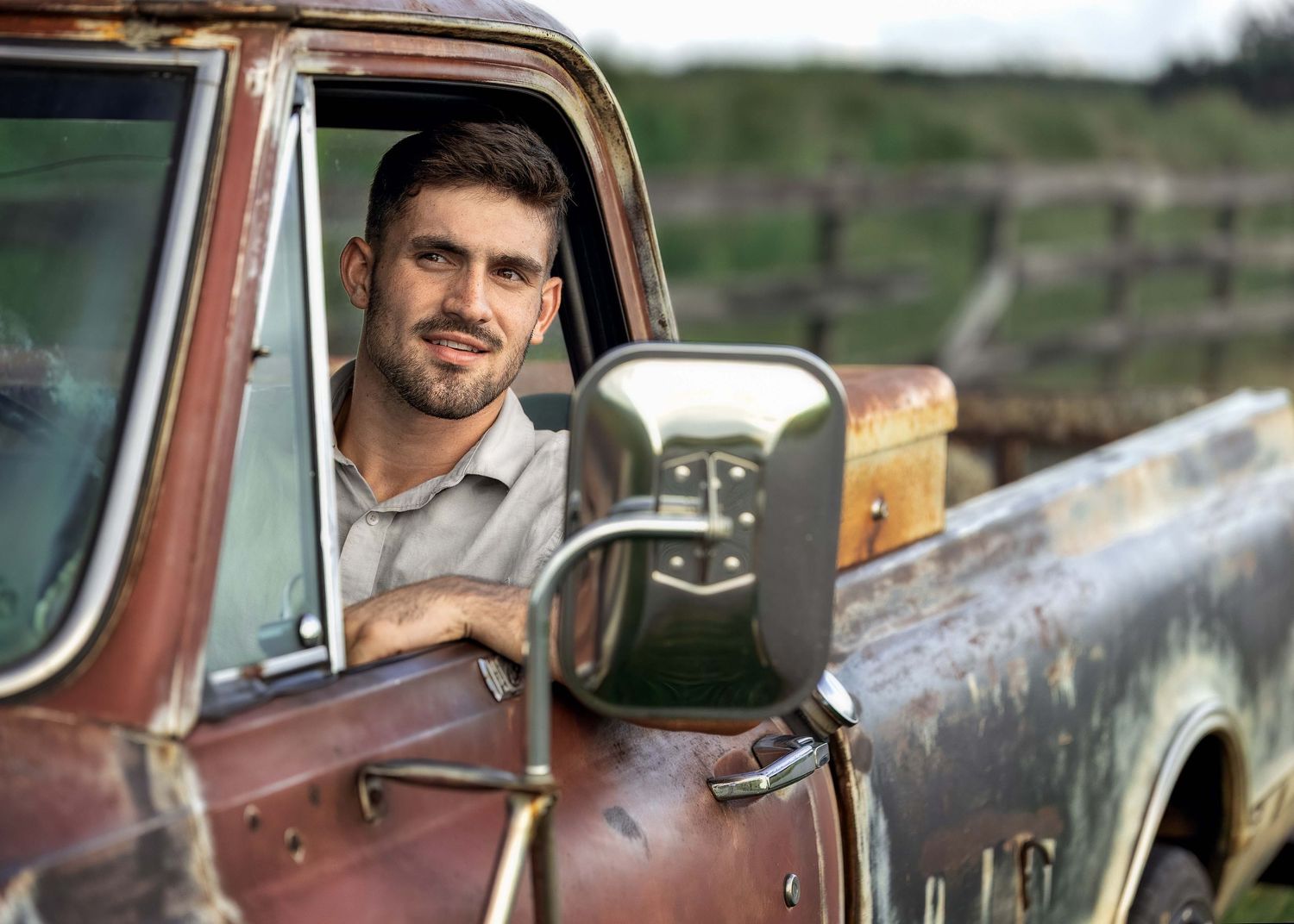 Young man with stubble smiling from rustic vintage truck window on a sunny day outdoors.