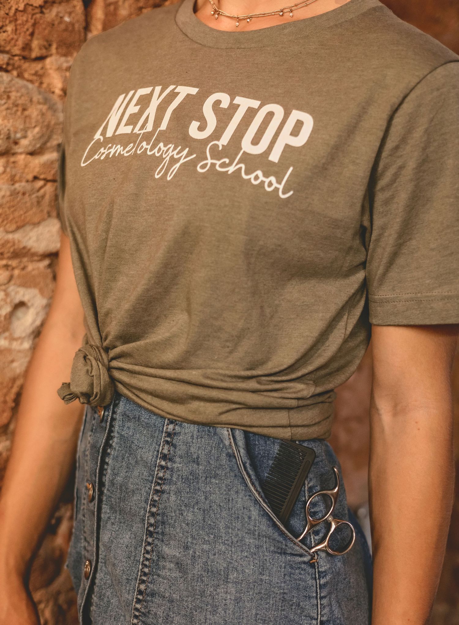 Olive green graphic tee reading Next Stop Cosmetology School, tied at waist, paired with denim skirt and scissors.