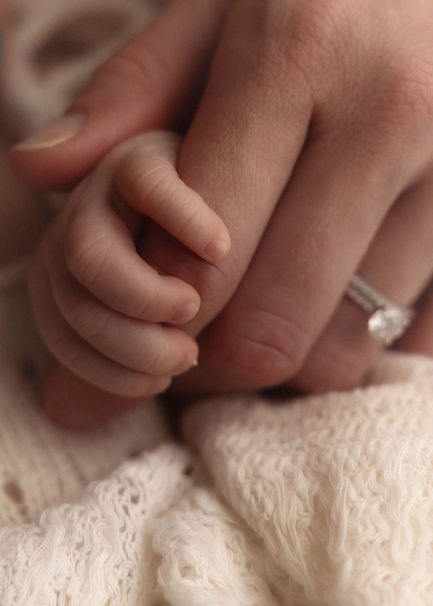 A newborn baby's tiny hand grasping an adult's finger, with a diamond engagement ring visible on the parent's hand.