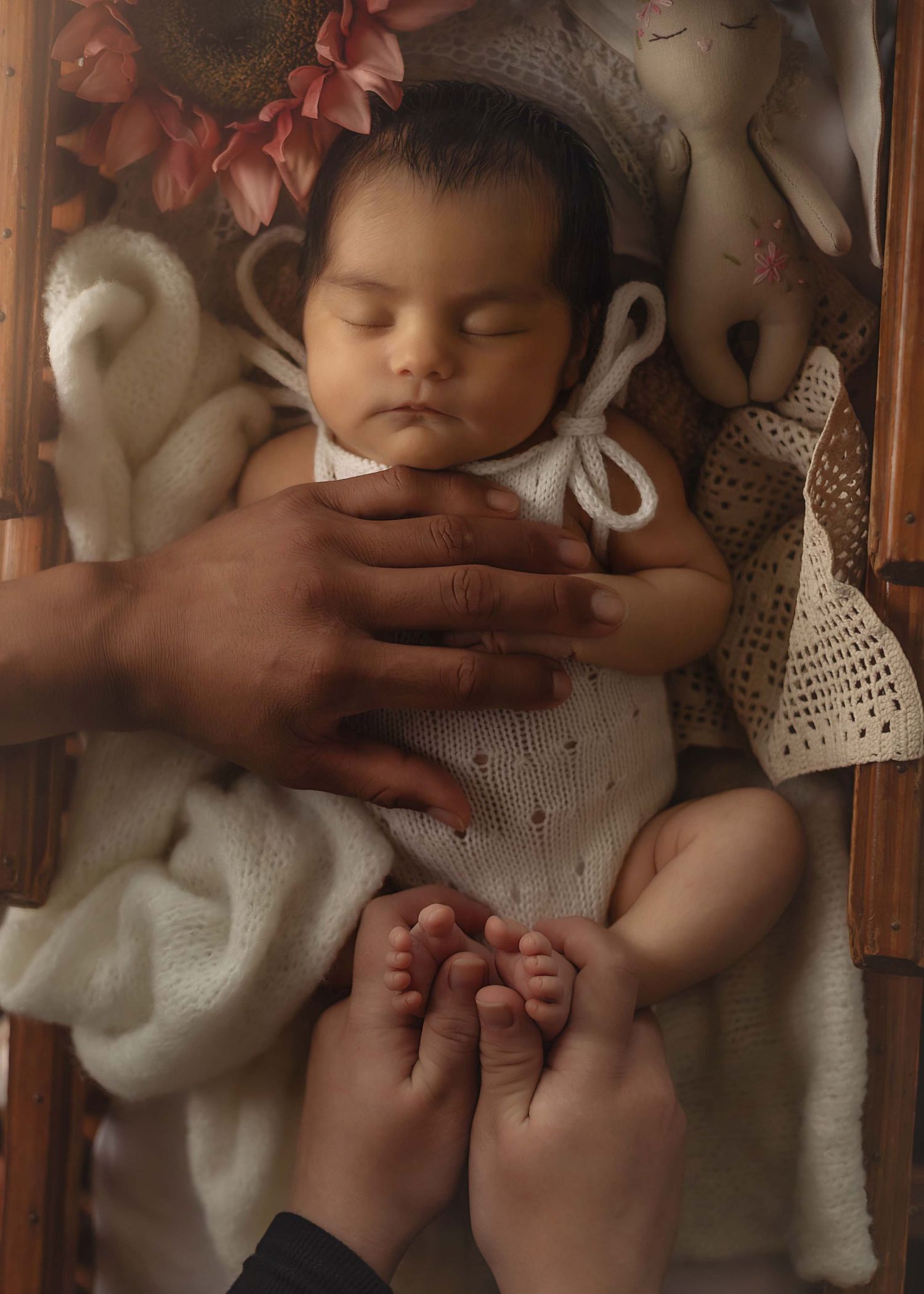 Newborn baby sleeping peacefully in a wooden crib, held gently by parents hands, wearing white knit outfit.
