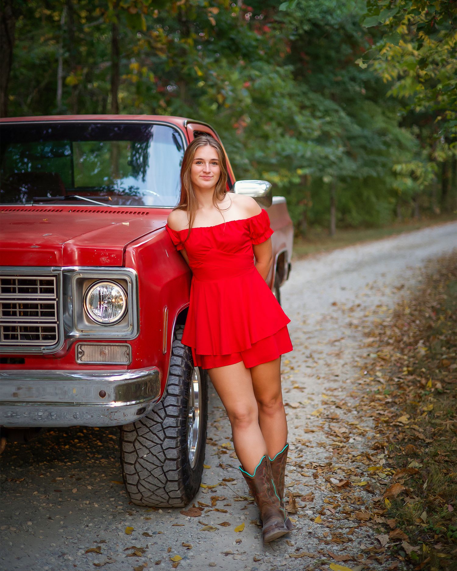 Young woman in red dress and cowboy boots leaning against a classic red truck on a scenic dirt road.