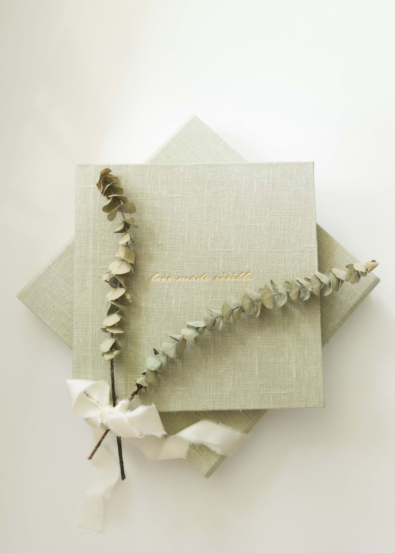 Sage green linen photo album with gold text, eucalyptus sprig, and white ribbon on white background.