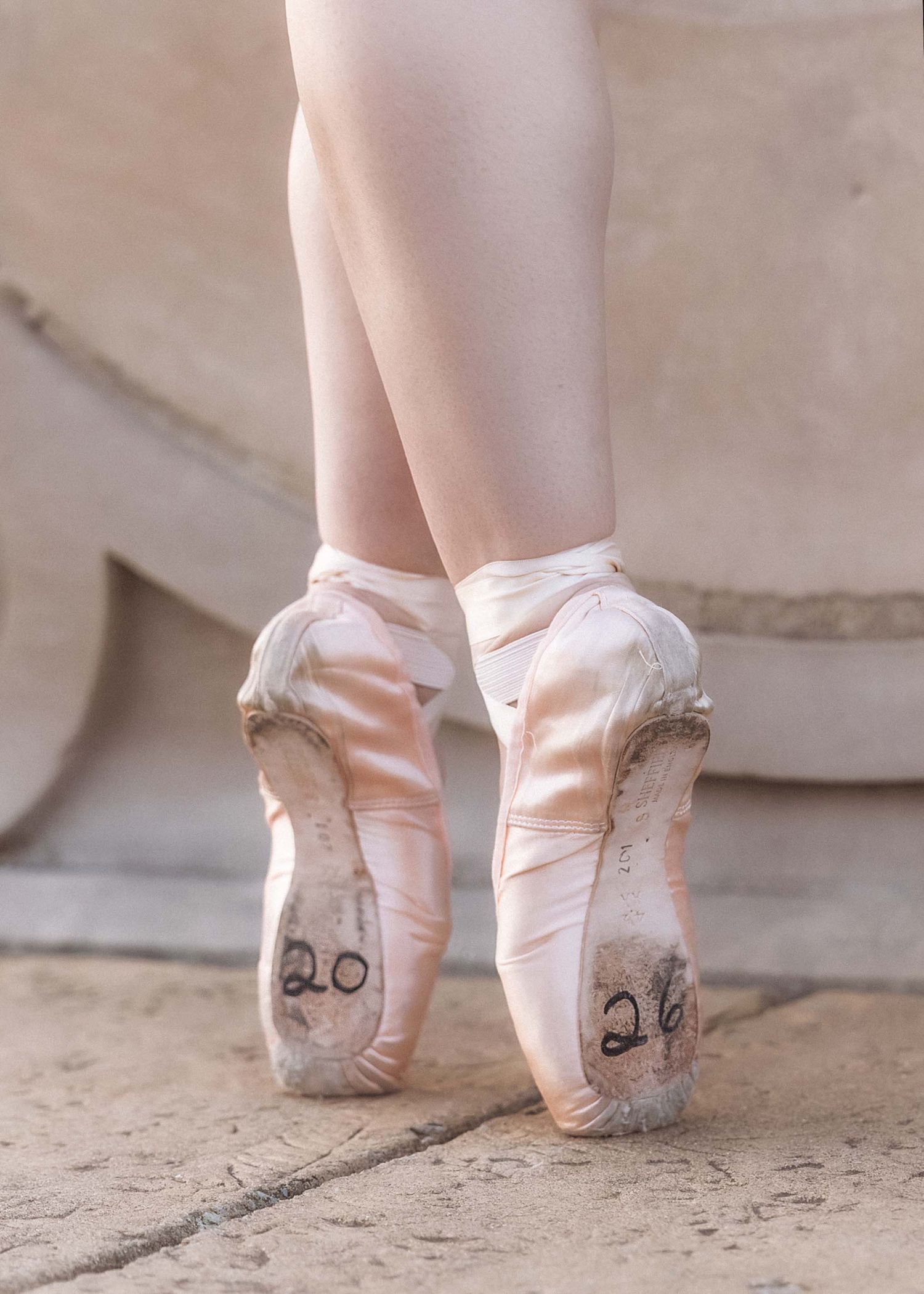 Worn pink ballet pointe shoes with ribbons tied around a dancer's ankles, numbered 20 and 26 on soles.