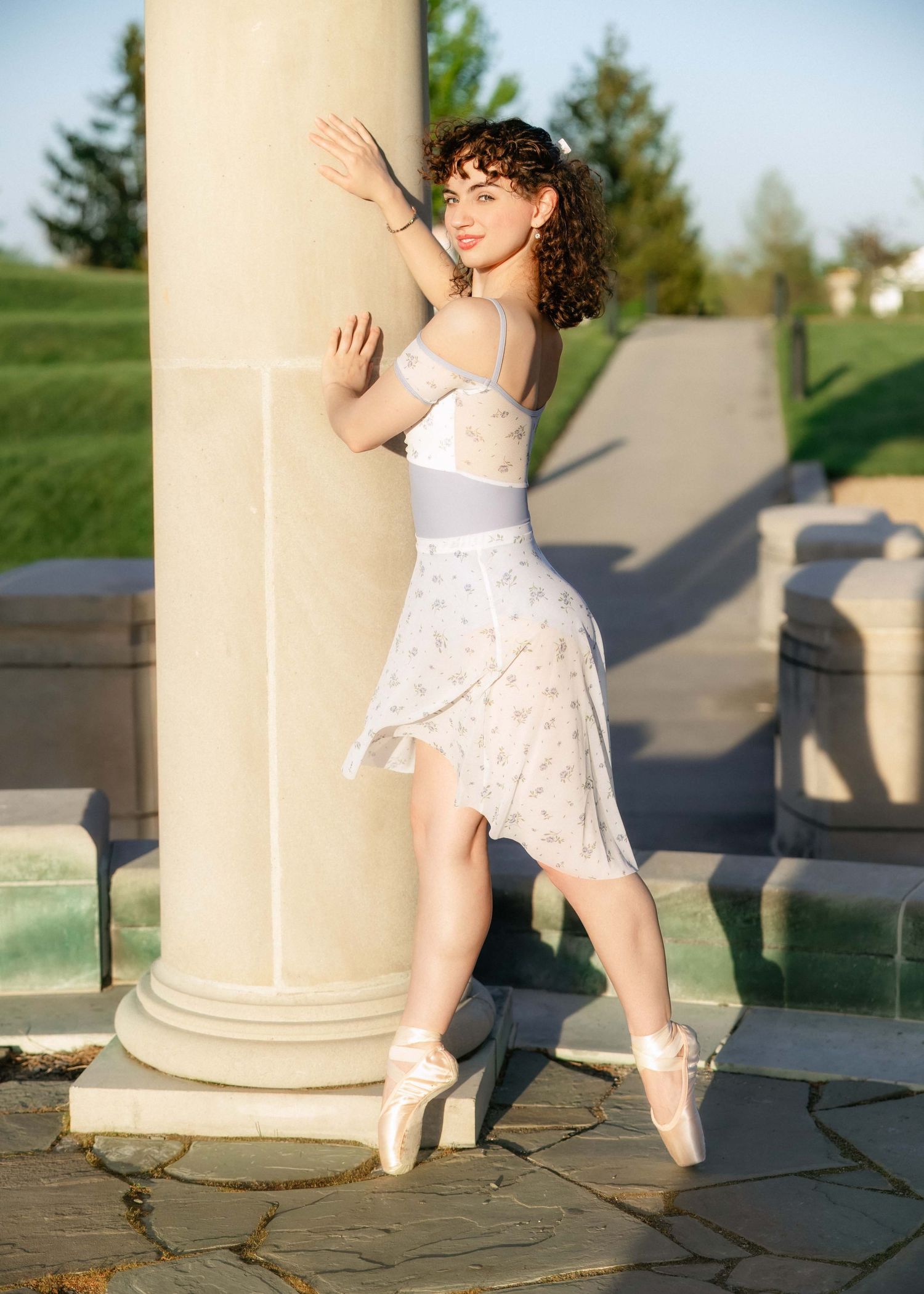 A young ballerina in a white floral skirt and pointe shoes poses gracefully beside a stone column outdoors.