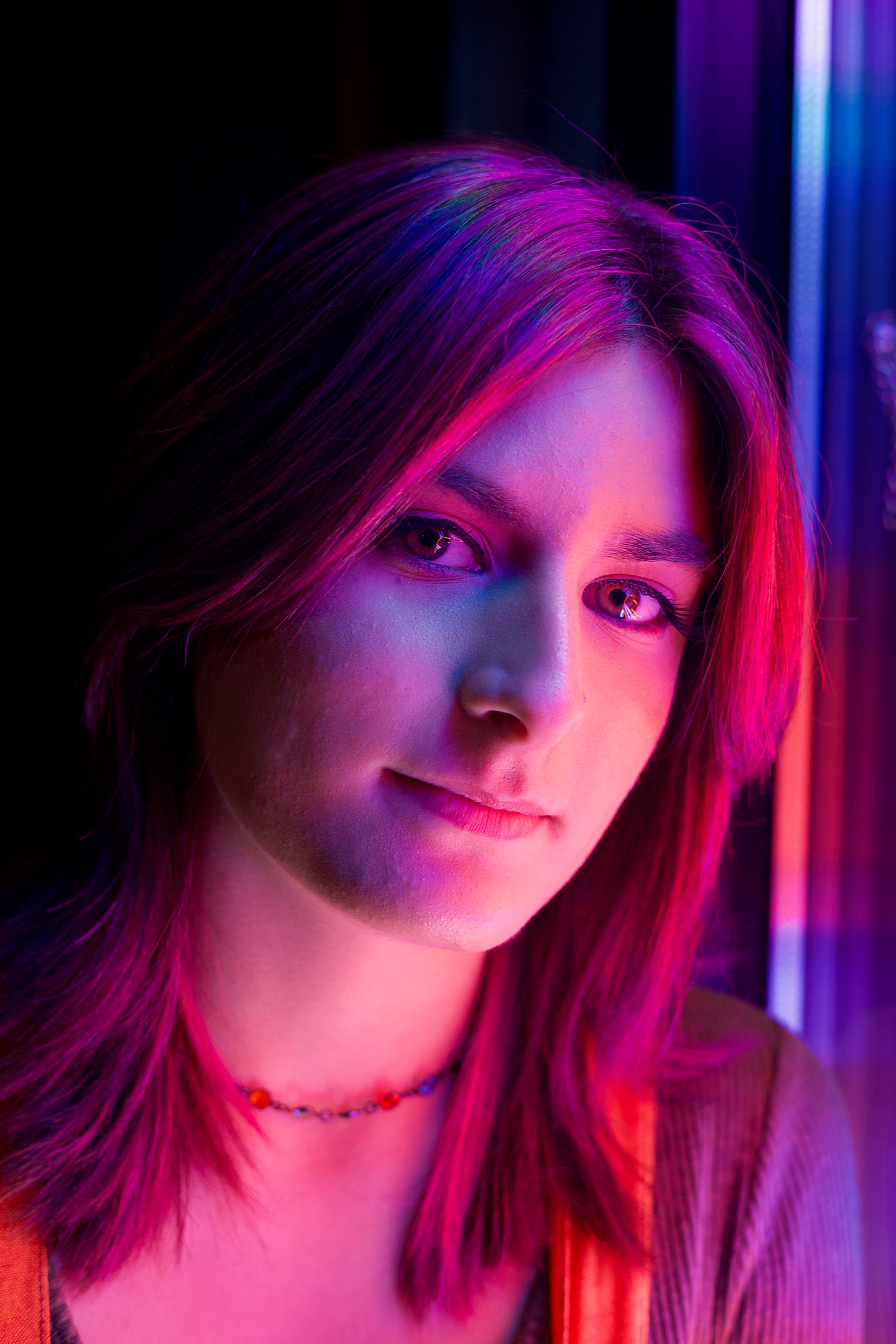 Young woman with red hair illuminated by vibrant pink and blue neon lights in a moody portrait.