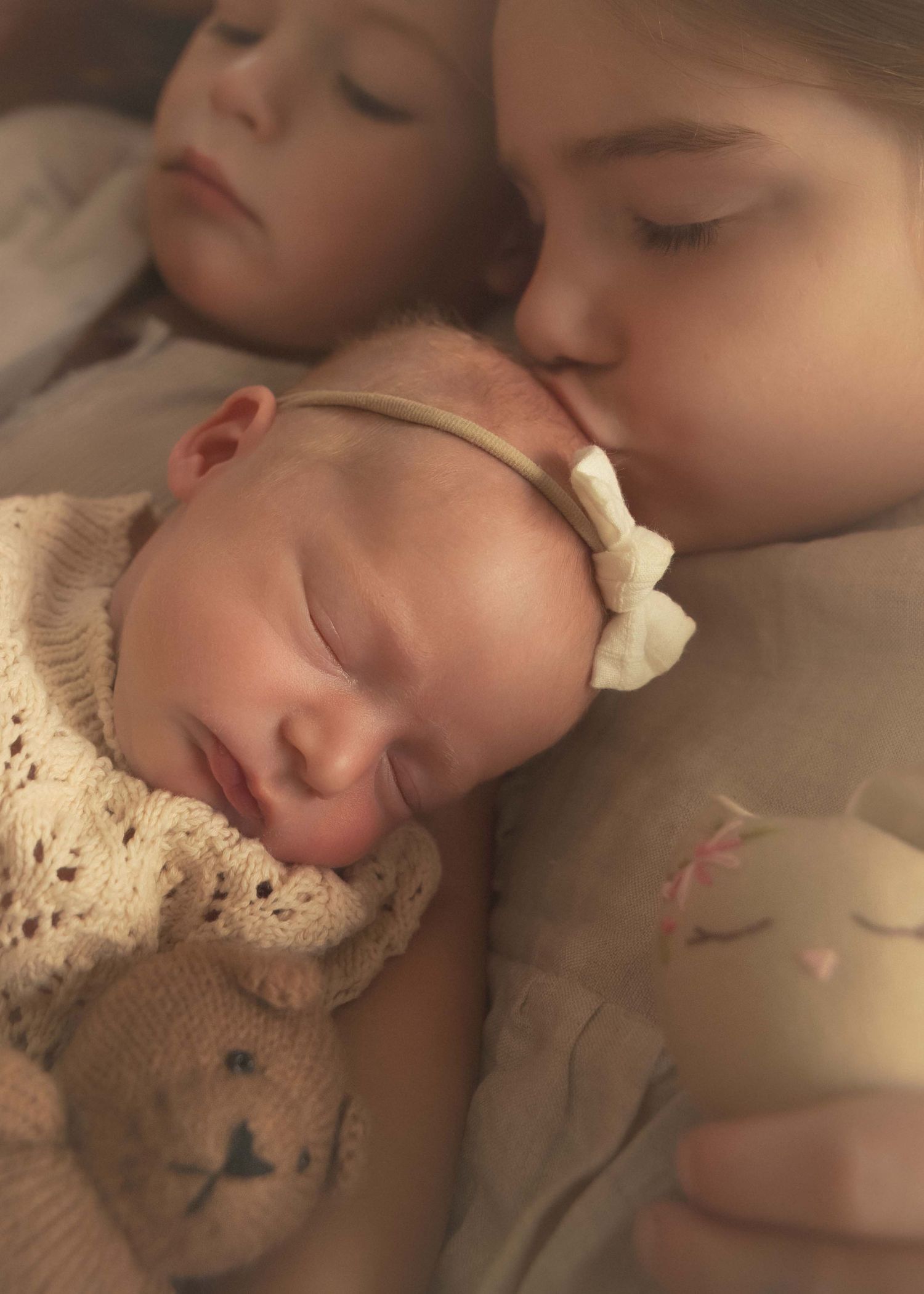 Older siblings sleeping peacefully with newborn baby wearing bow headband and holding teddy bear.