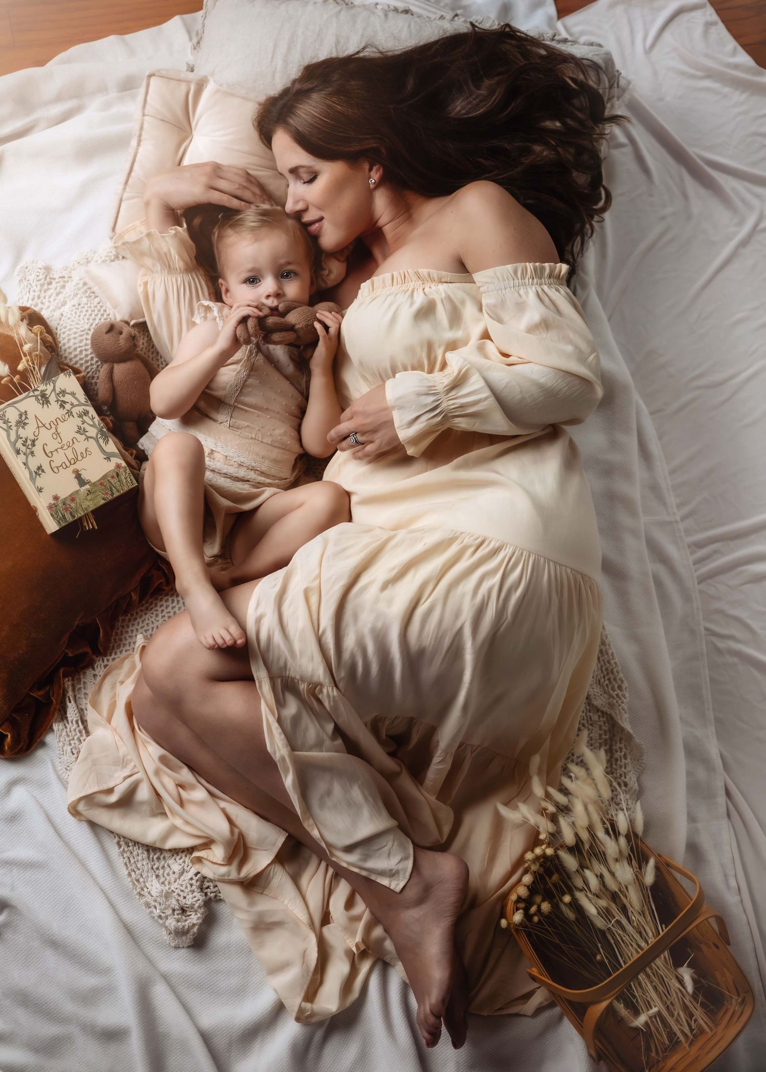 Mother and baby lying together in soft cream colored clothing on white bedding.