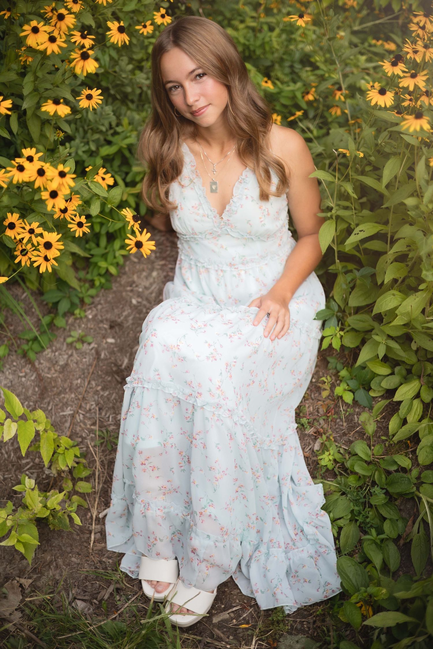 Young woman in floral maxi dress sitting among yellow black-eyed Susan flowers in a garden.