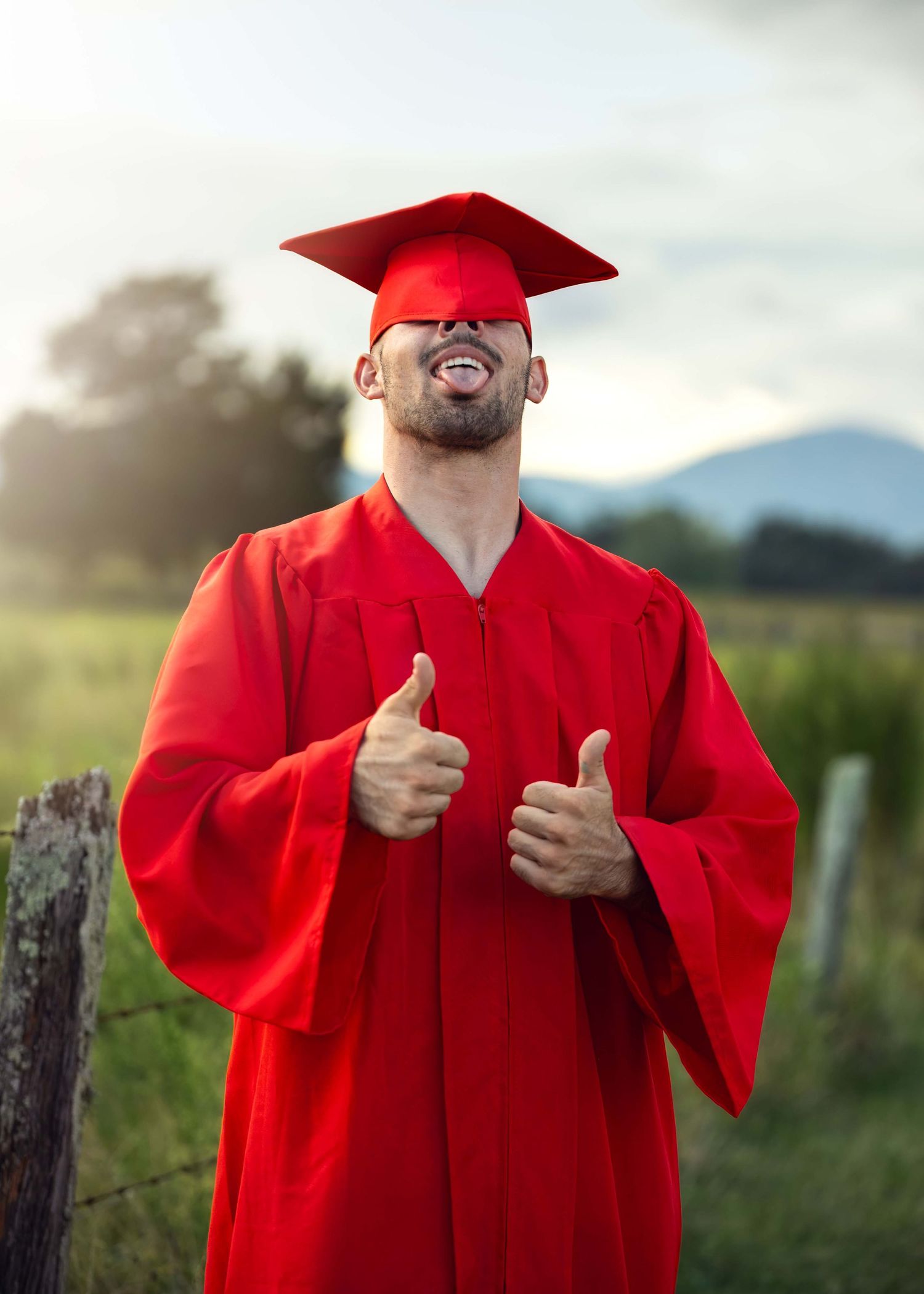 Man in red graduation cap and gown giving two thumbs up outdoors with green fields and mountains behind.