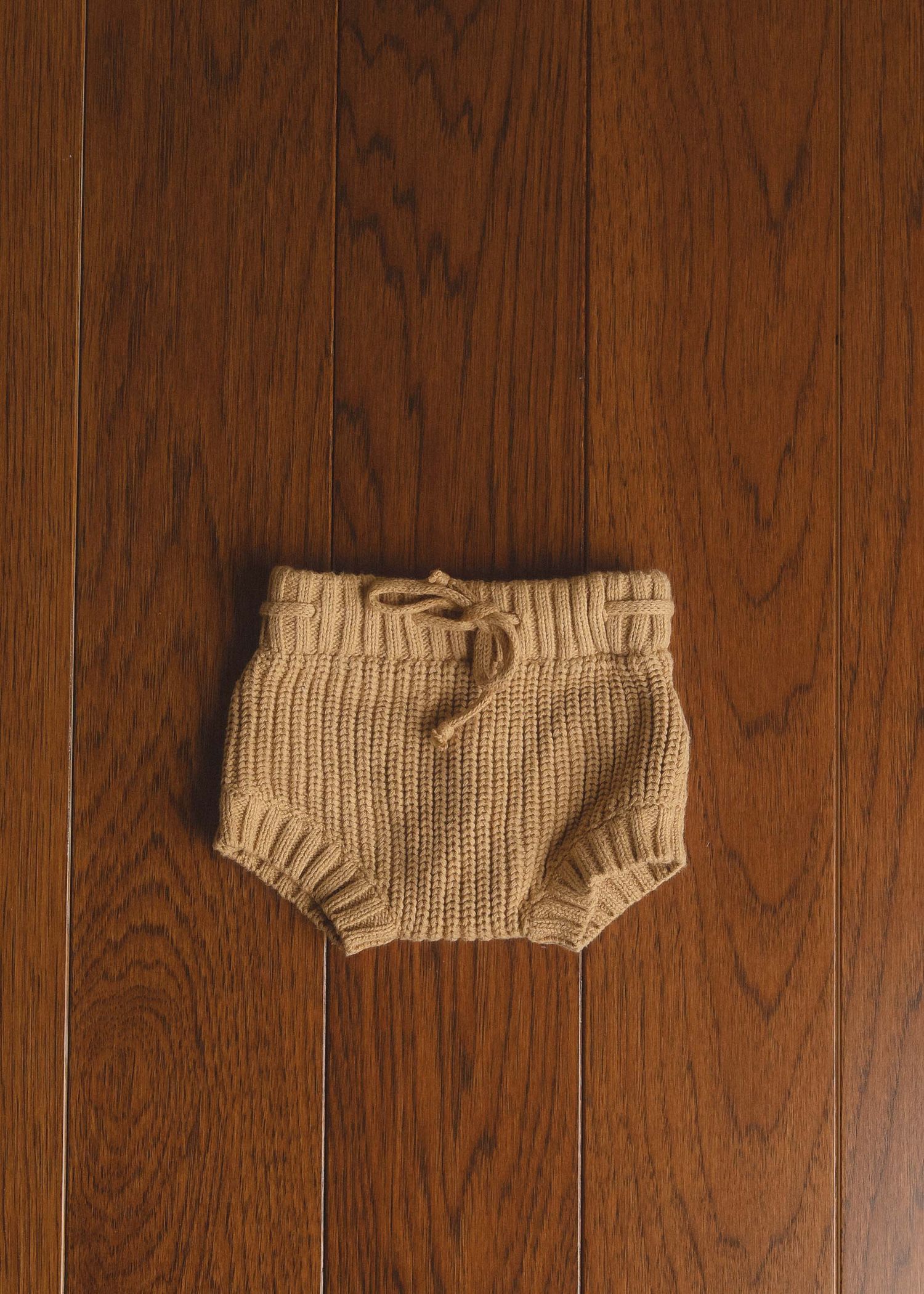 Tan knit baby bloomers with drawstring waist displayed on dark wood floor.