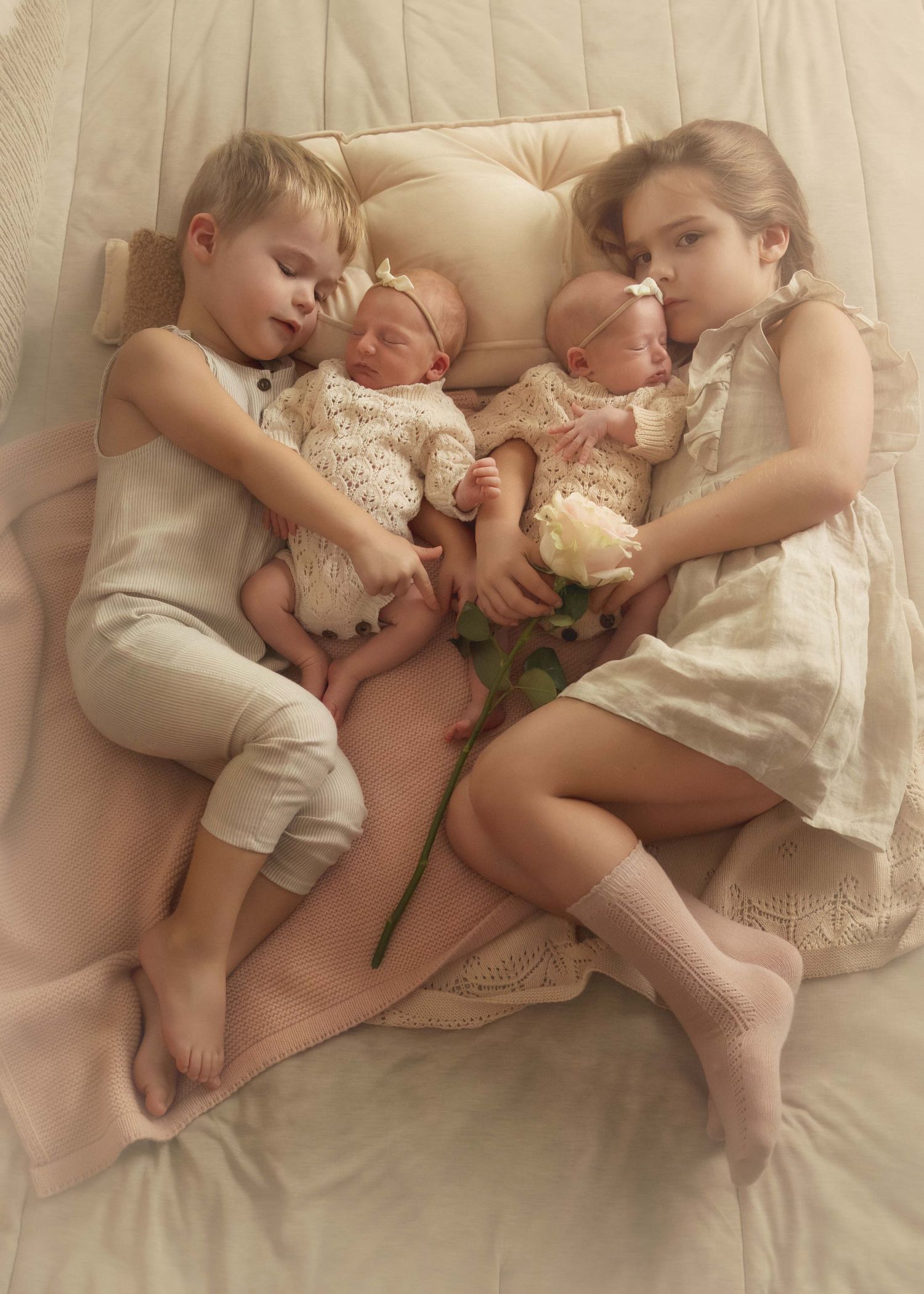 Two older siblings cuddle newborn twins in lace outfits on a soft bed, holding a white rose.