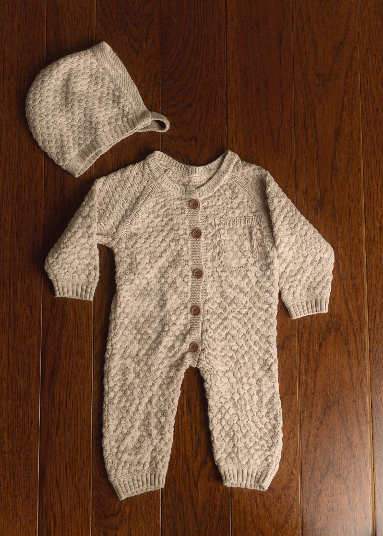Cream knit baby romper with wooden buttons and matching bonnet hat on wood floor.