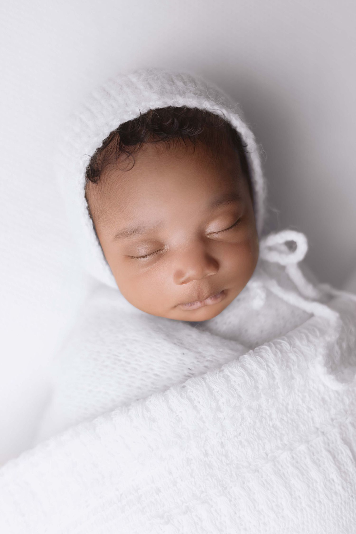 Sleeping newborn baby wrapped in white knit blanket and bonnet on white background.
