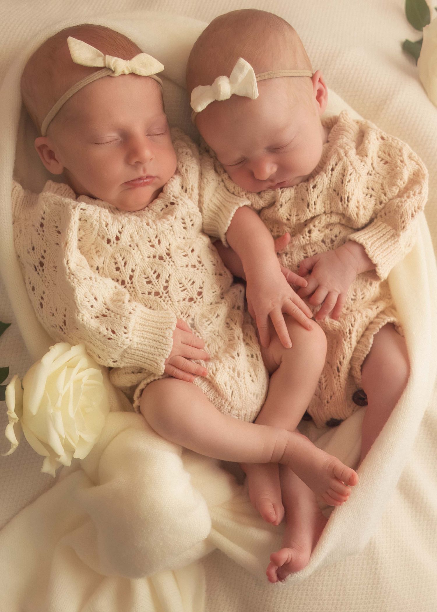Two sleeping newborn twins in cream knit outfits and bow headbands cuddle together on a white blanket.