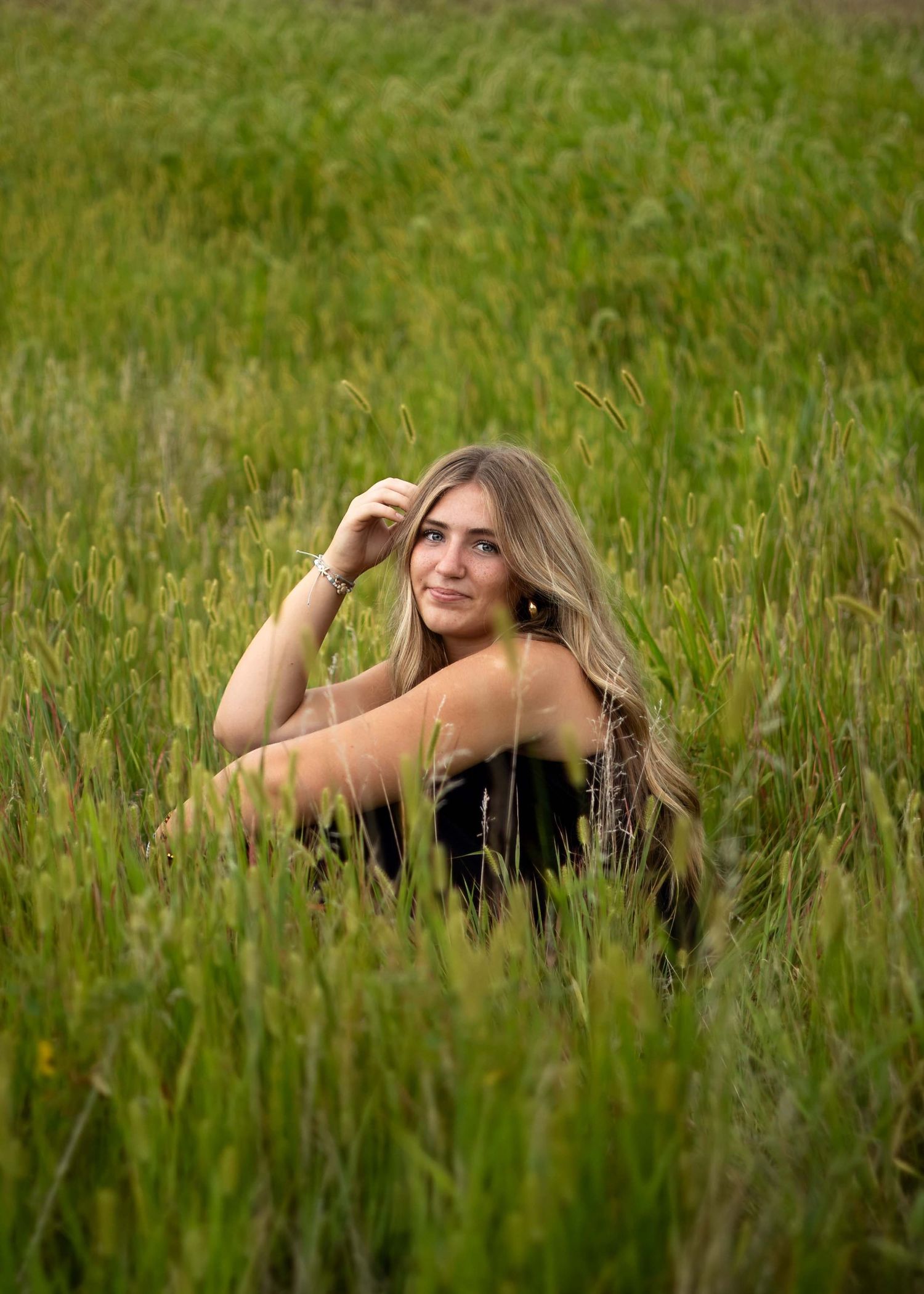 Young woman with long blonde hair sitting in a lush green field, posing gracefully outdoors.