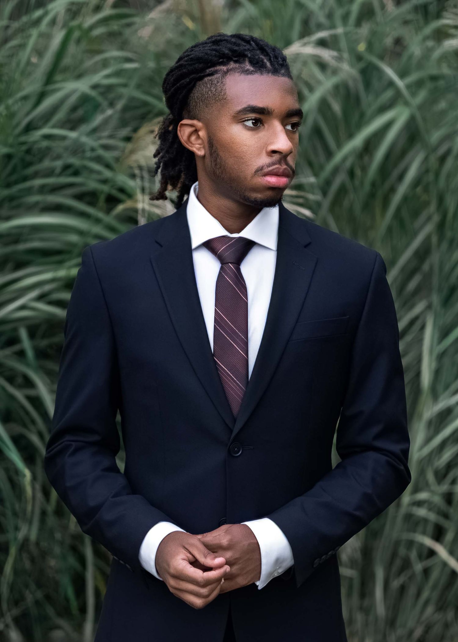 Young Black man in black suit and striped tie standing outdoors near tall grass.