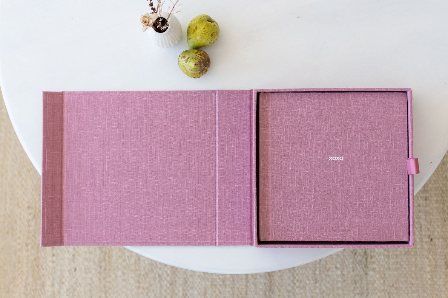 Pink linen photo album box set displayed open on a white round table with decorative green spheres.