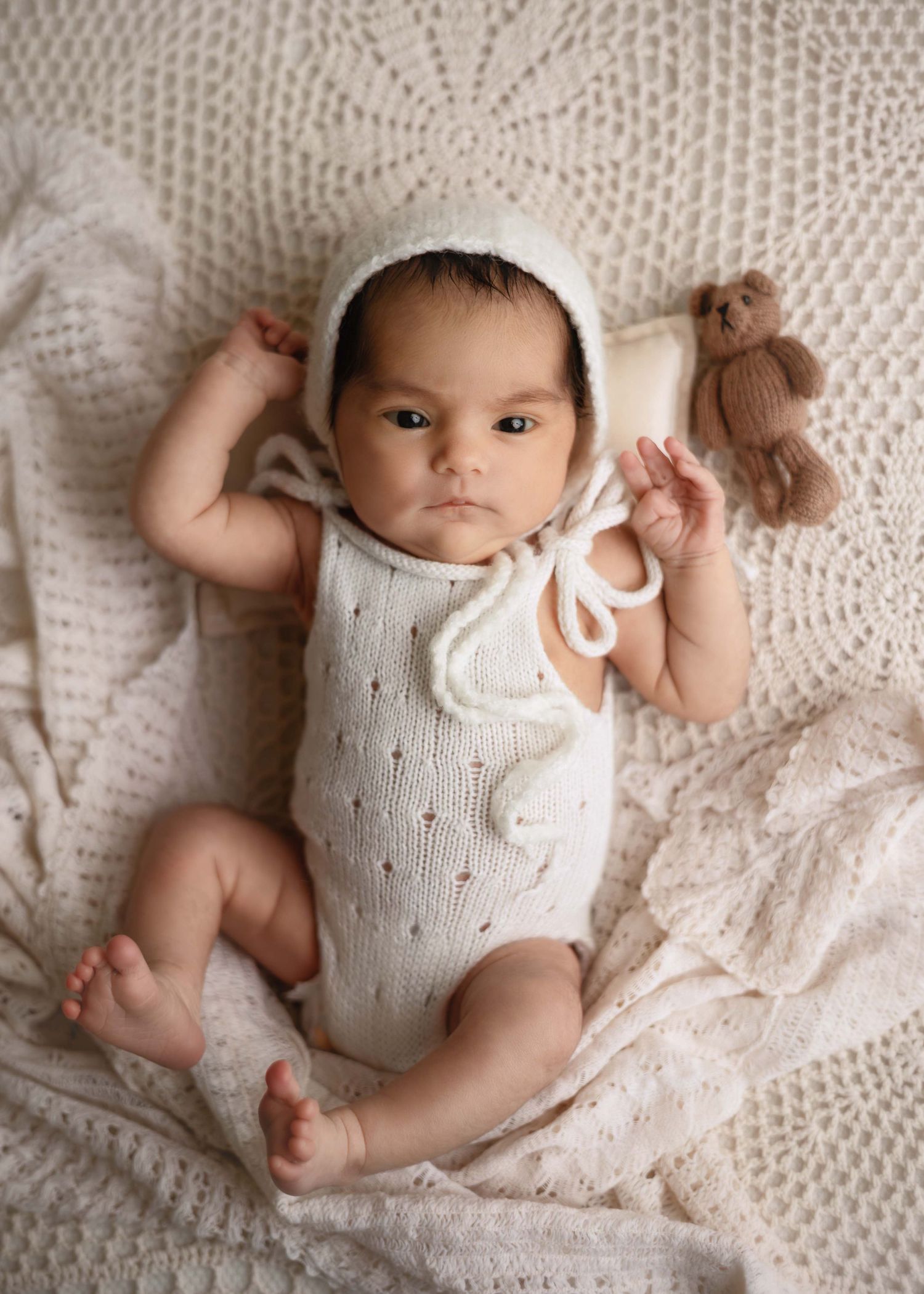 Newborn baby in cream knit romper and bonnet lying on white crochet blanket holding a small teddy bear.