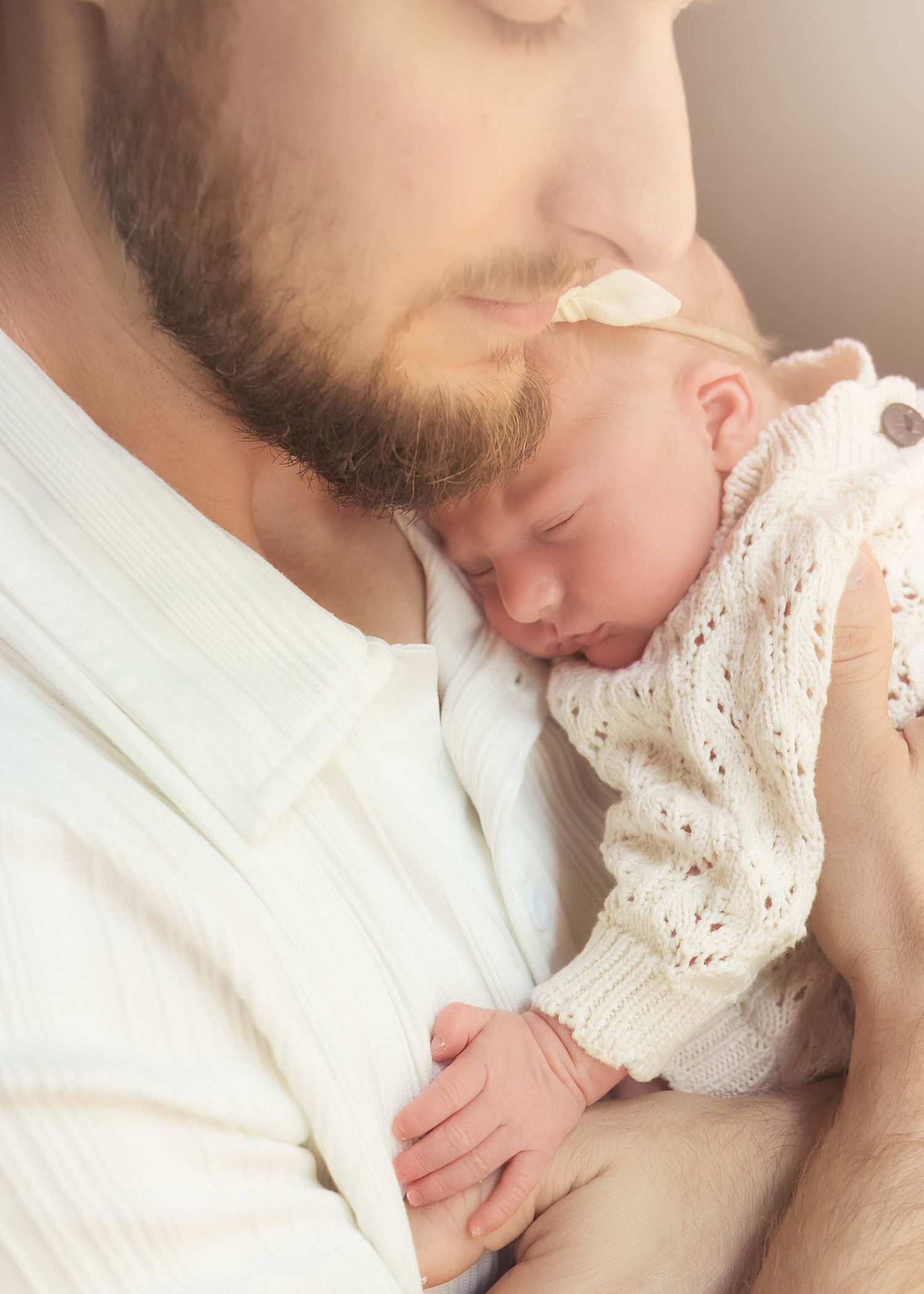 A bearded father tenderly holds his sleeping newborn baby dressed in a white knit outfit close to his chest.