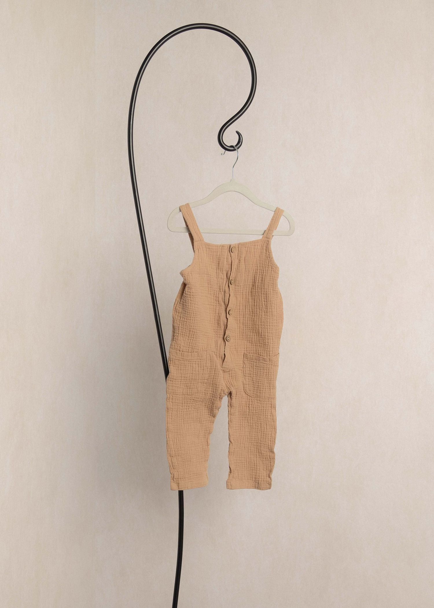 Beige muslin baby romper with button front detail, hanging on a decorative black hook against a neutral wall.