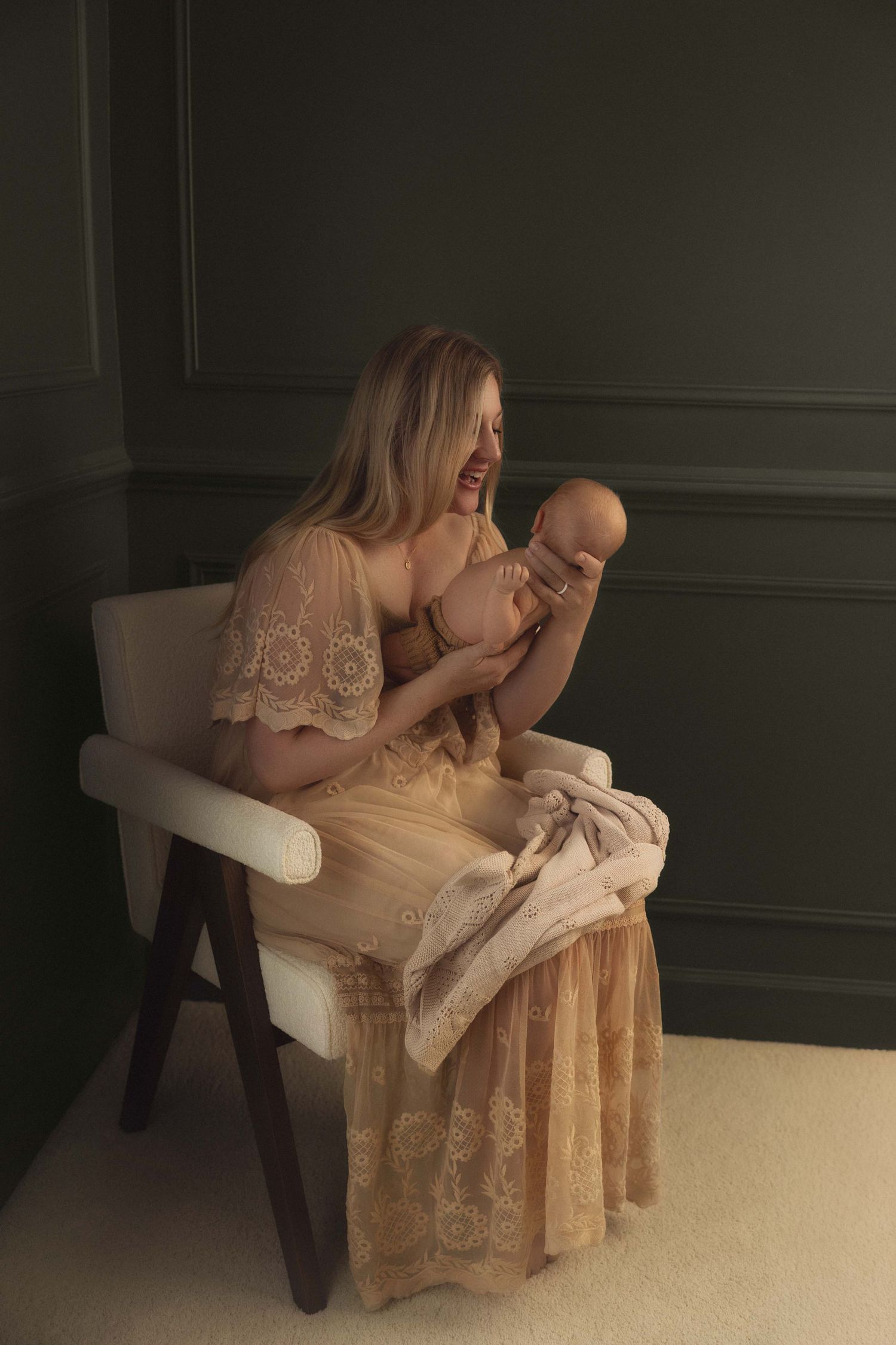 A mother in a lace gown sits on a white chair, lovingly cradling her newborn baby in a softly lit room.