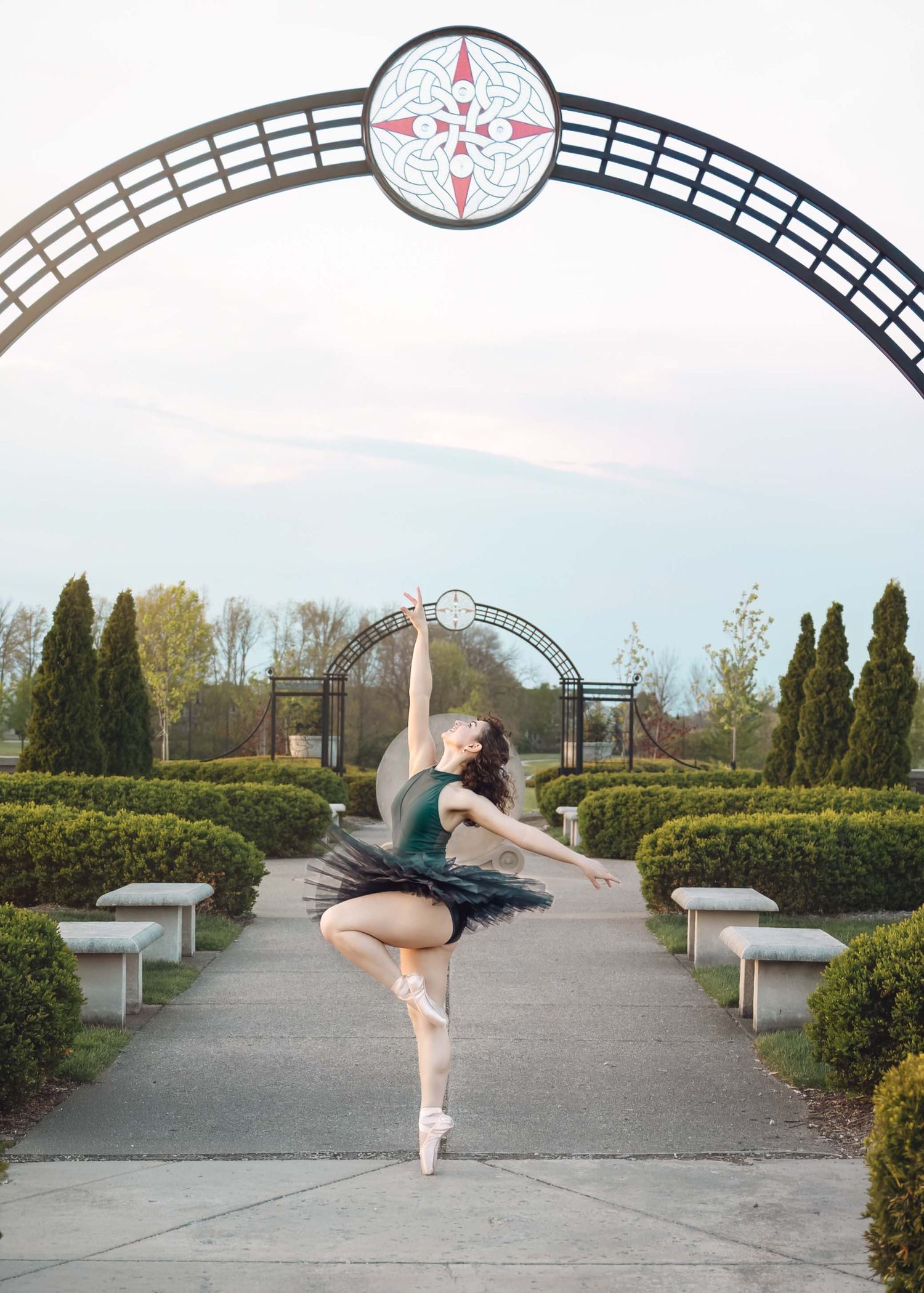 A ballerina in a black tutu poses en pointe under a decorative arch in a formal garden.