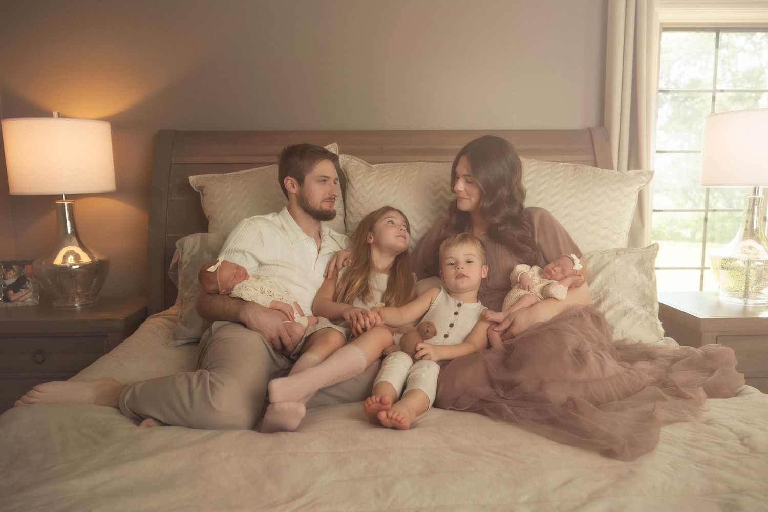 A family of six sits together on a bed, with two young children between their parents in a warmly lit bedroom.