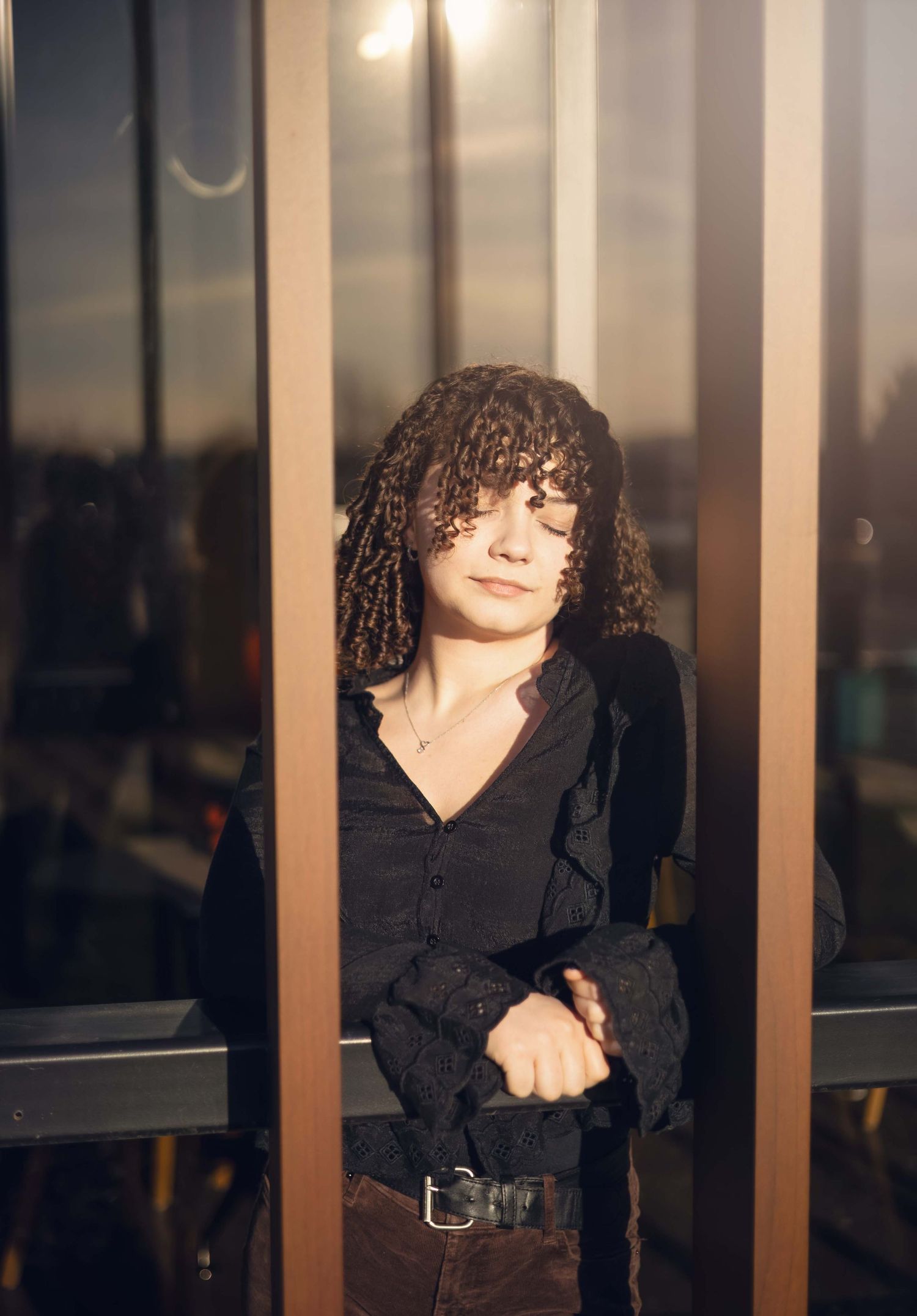 Young woman with curly hair leaning on metal railing in warm golden hour sunlight.