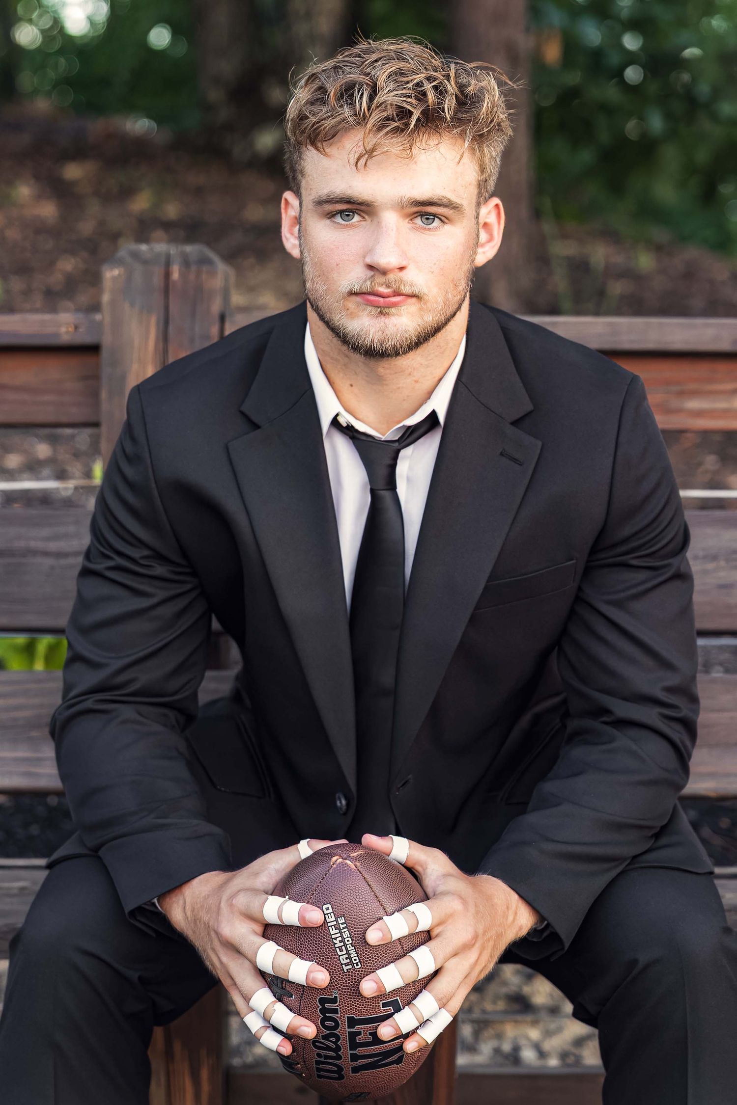 Young man in black suit holding NFL Wilson football while sitting on a bench outdoors.