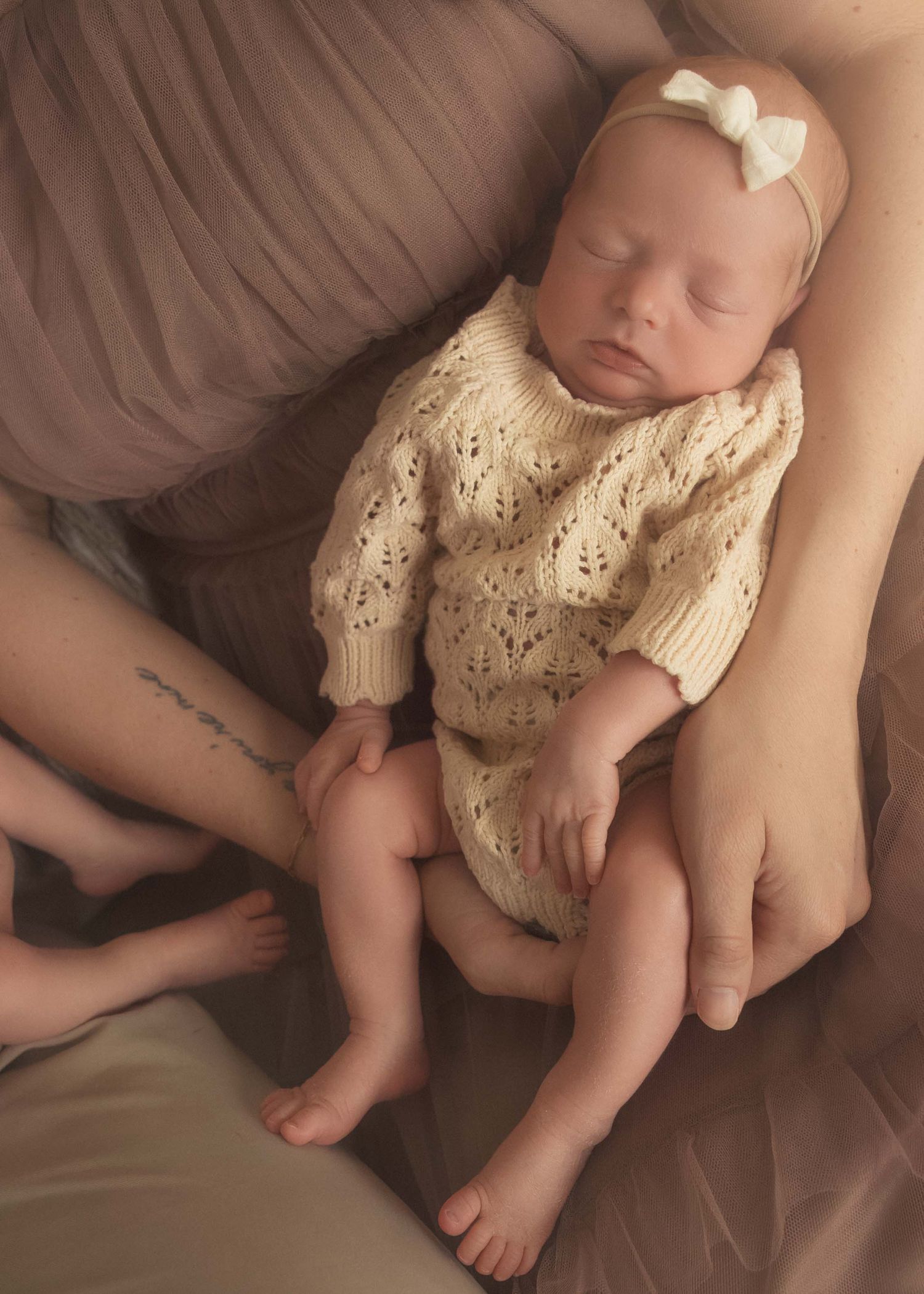 Sleeping newborn baby in cream knit romper with white bow headband, cradled in parents arms on soft pink backdrop.
