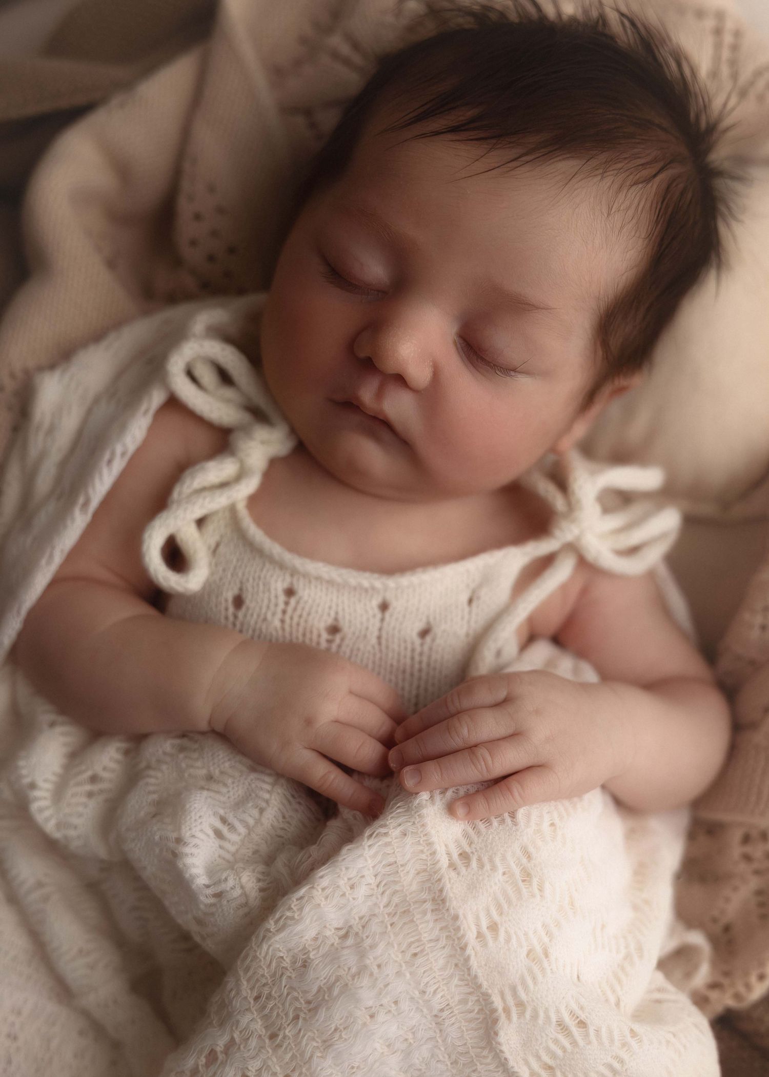 Peaceful newborn baby sleeping in a white knit romper, wrapped in soft cream blankets.