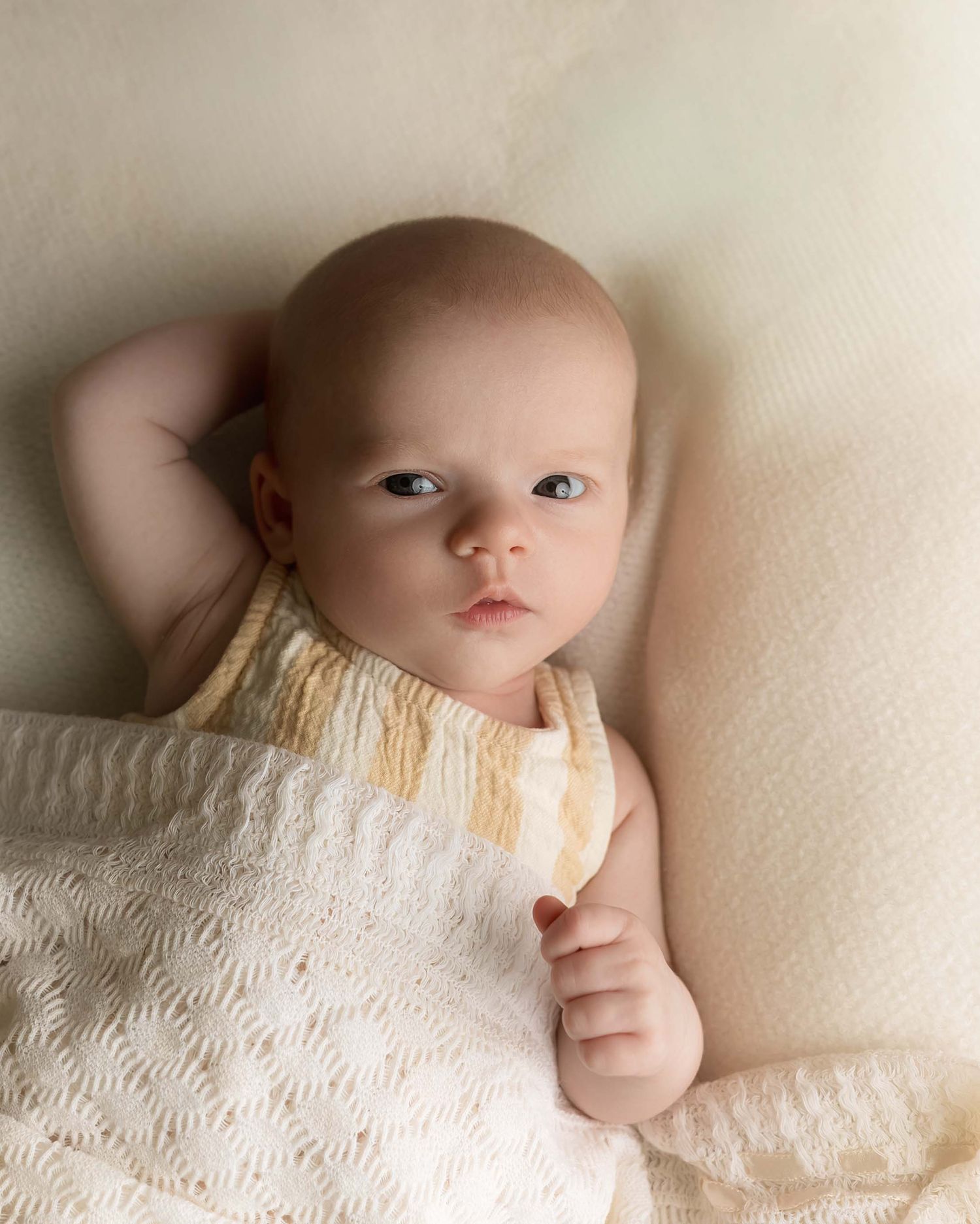 A serene baby lying on a cream knit blanket, wearing a white lace outfit, gazing softly with bright blue eyes.