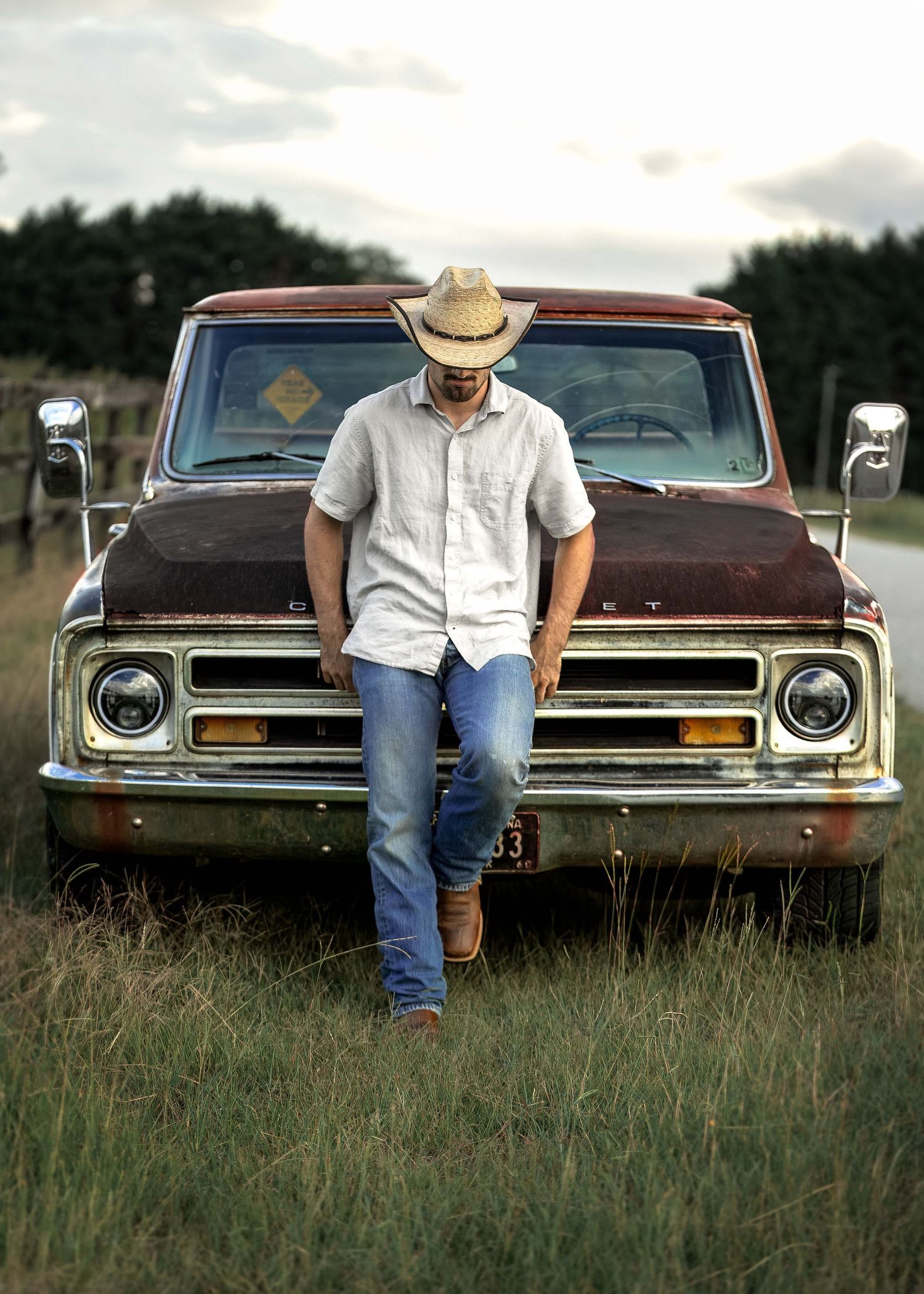 Man in cowboy hat and jeans leaning against a rustic vintage Chevy pickup truck in a rural field at dusk.