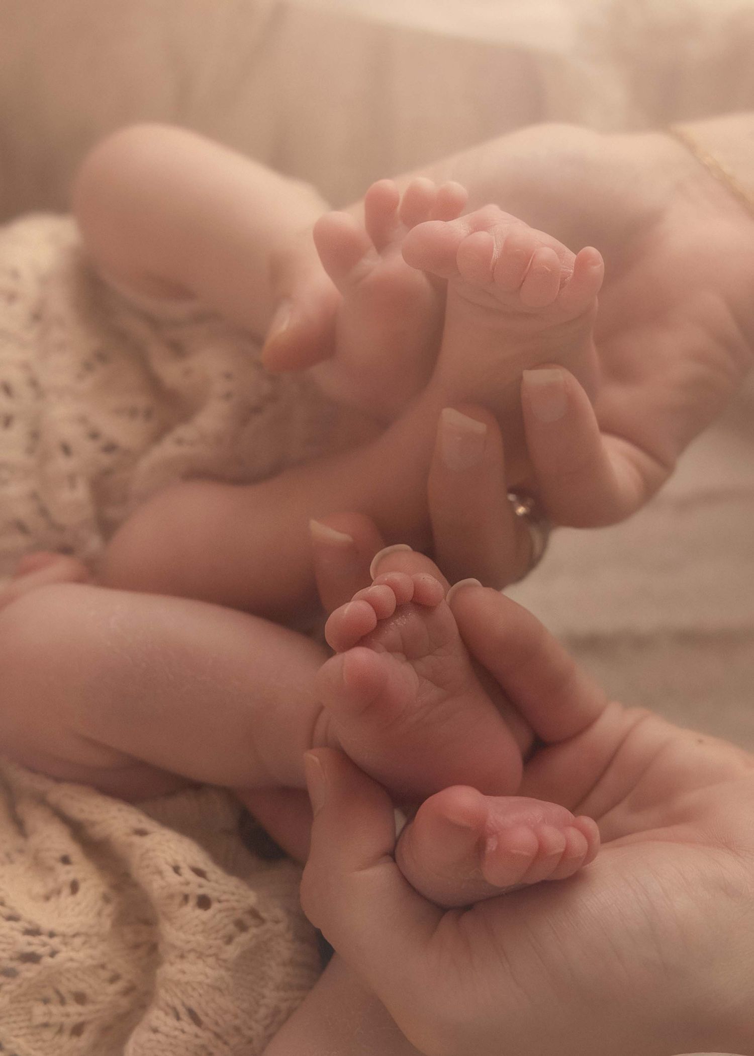Adult hands gently cradling two newborn babies tiny feet together, soft warm light highlighting delicate skin tones.