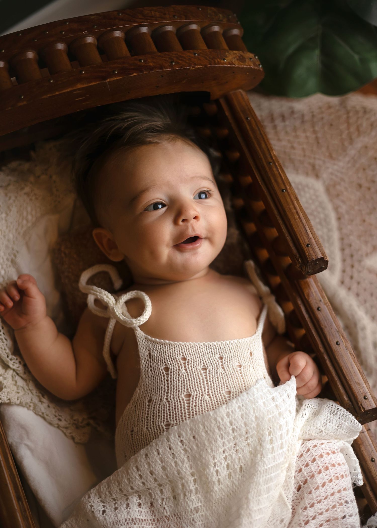Smiling baby in a white knit dress lying in a vintage wooden cradle with lace bedding.