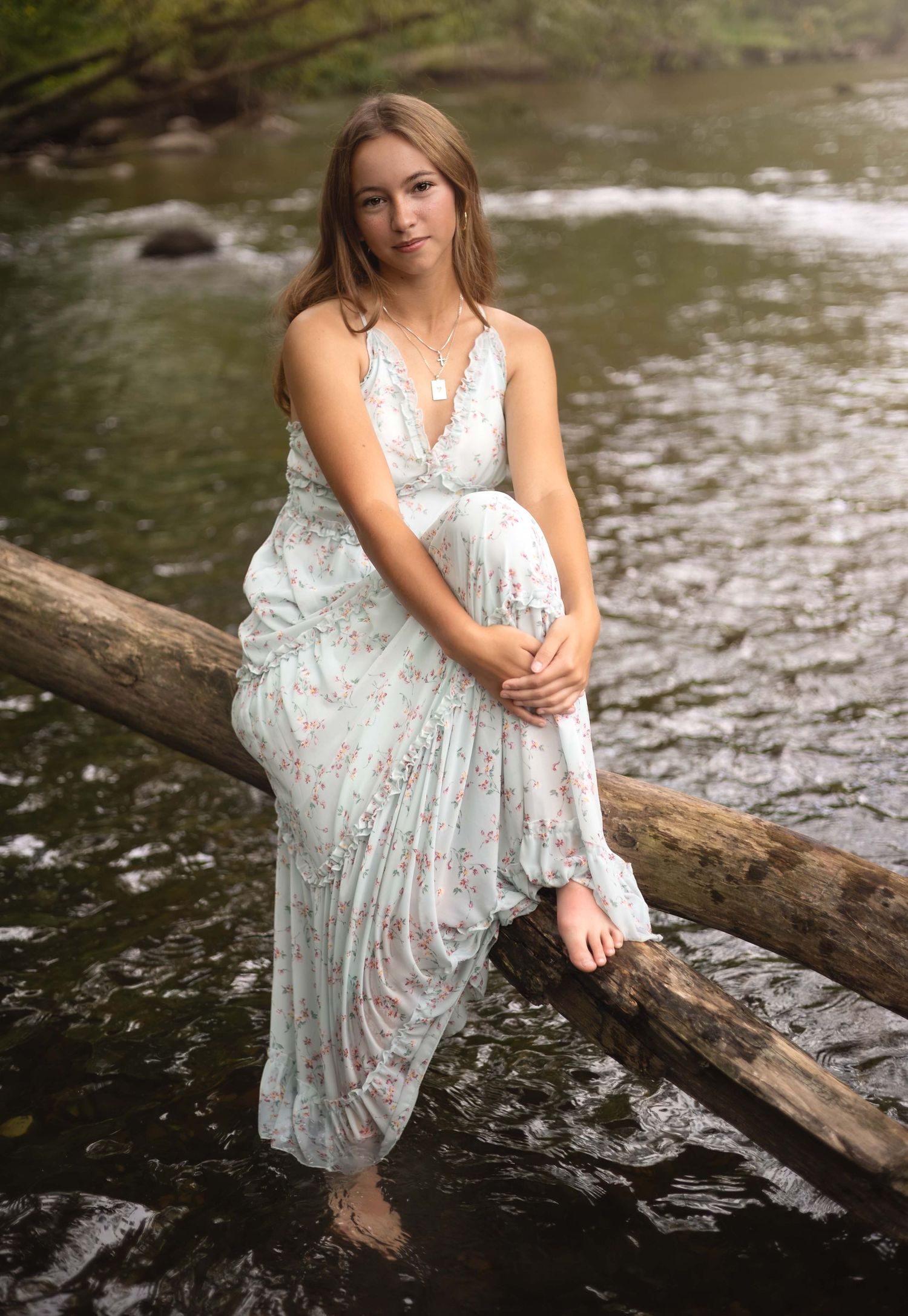 Young woman in floral maxi dress sitting on log over creek, barefoot in nature setting.