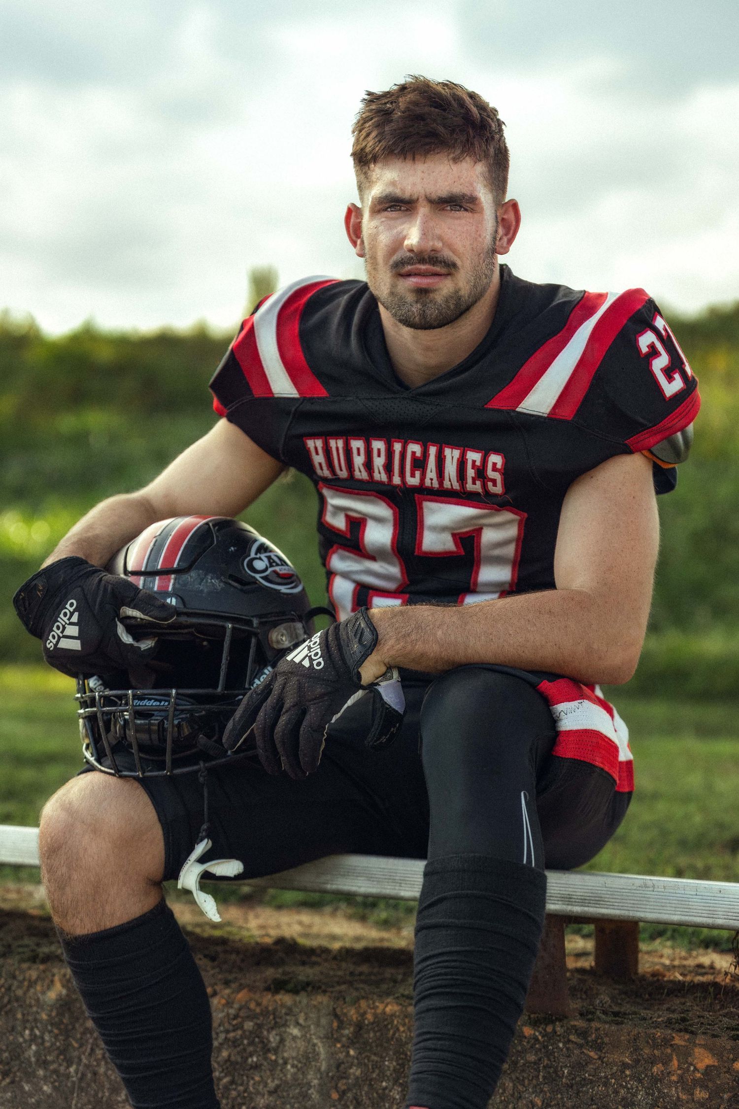 A Hurricanes football player in black and red uniform #27 sits on a bench holding his helmet outdoors.