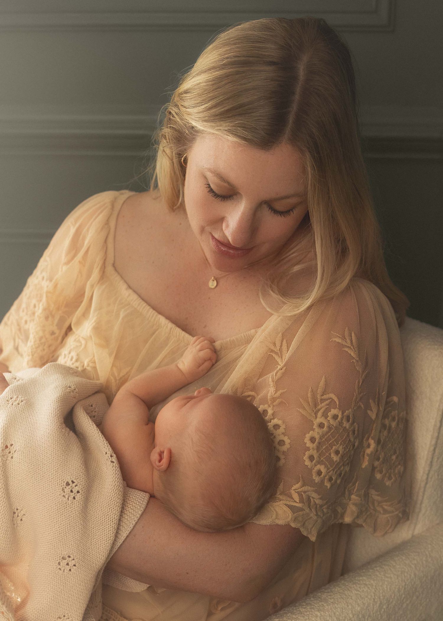 A mother in a cream lace dress tenderly breastfeeds her newborn baby, wrapped in a soft blanket.