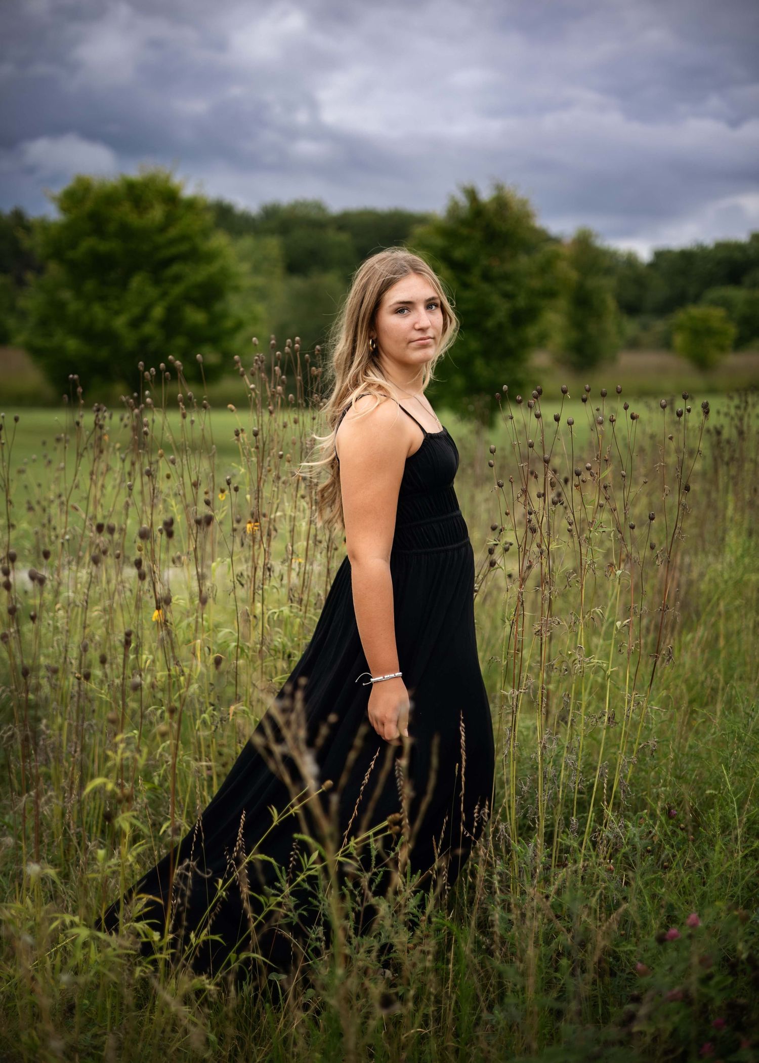 Young woman in elegant black maxi dress standing gracefully in a lush green meadow under dramatic cloudy skies.