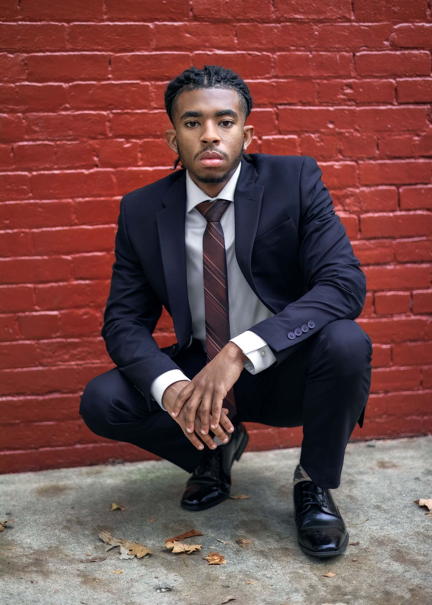 Young Black man in navy suit and burgundy tie crouching confidently against red brick wall outdoors.