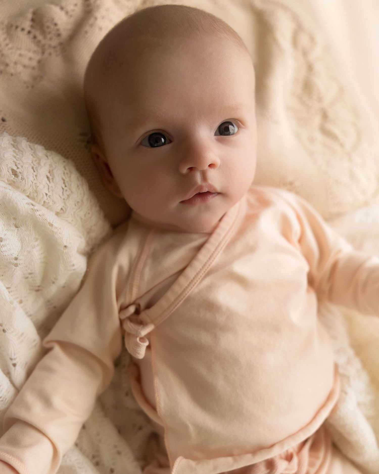 Adorable baby in a peach wrap onesie lying on a cream knit blanket, looking up with bright curious eyes.
