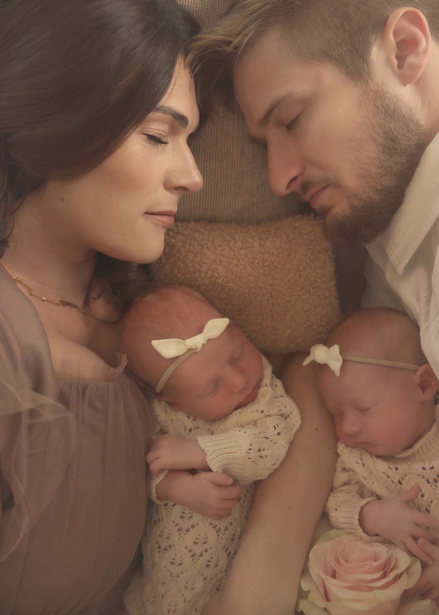 A couple with closed eyes lovingly hold their twin newborns wrapped in soft blankets with delicate bow headbands.