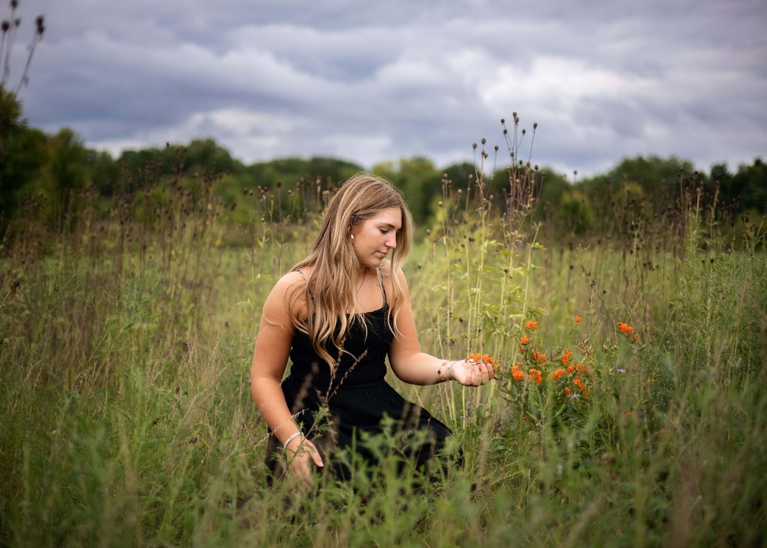 A young woman in a black dress kneels in a wildflower meadow, gently touching orange flowers under a cloudy sky.