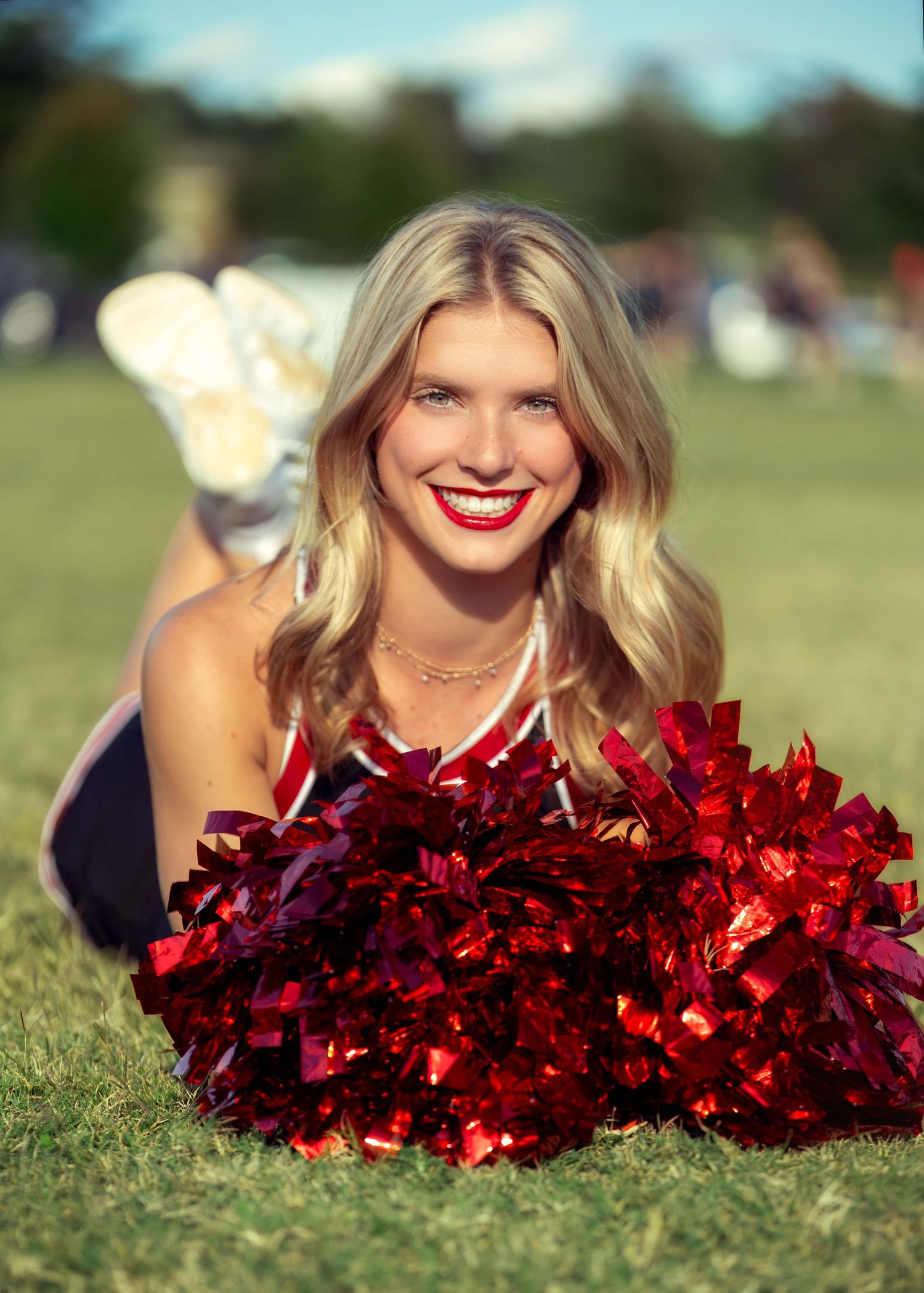 Smiling blonde cheerleader lying on grass holding red metallic pom-poms in a red uniform on a sunny day.