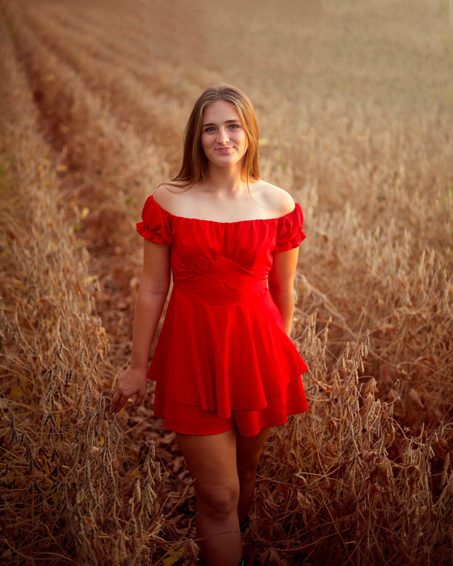 Young woman in red off-shoulder dress standing in a golden field, smiling warmly at the camera.