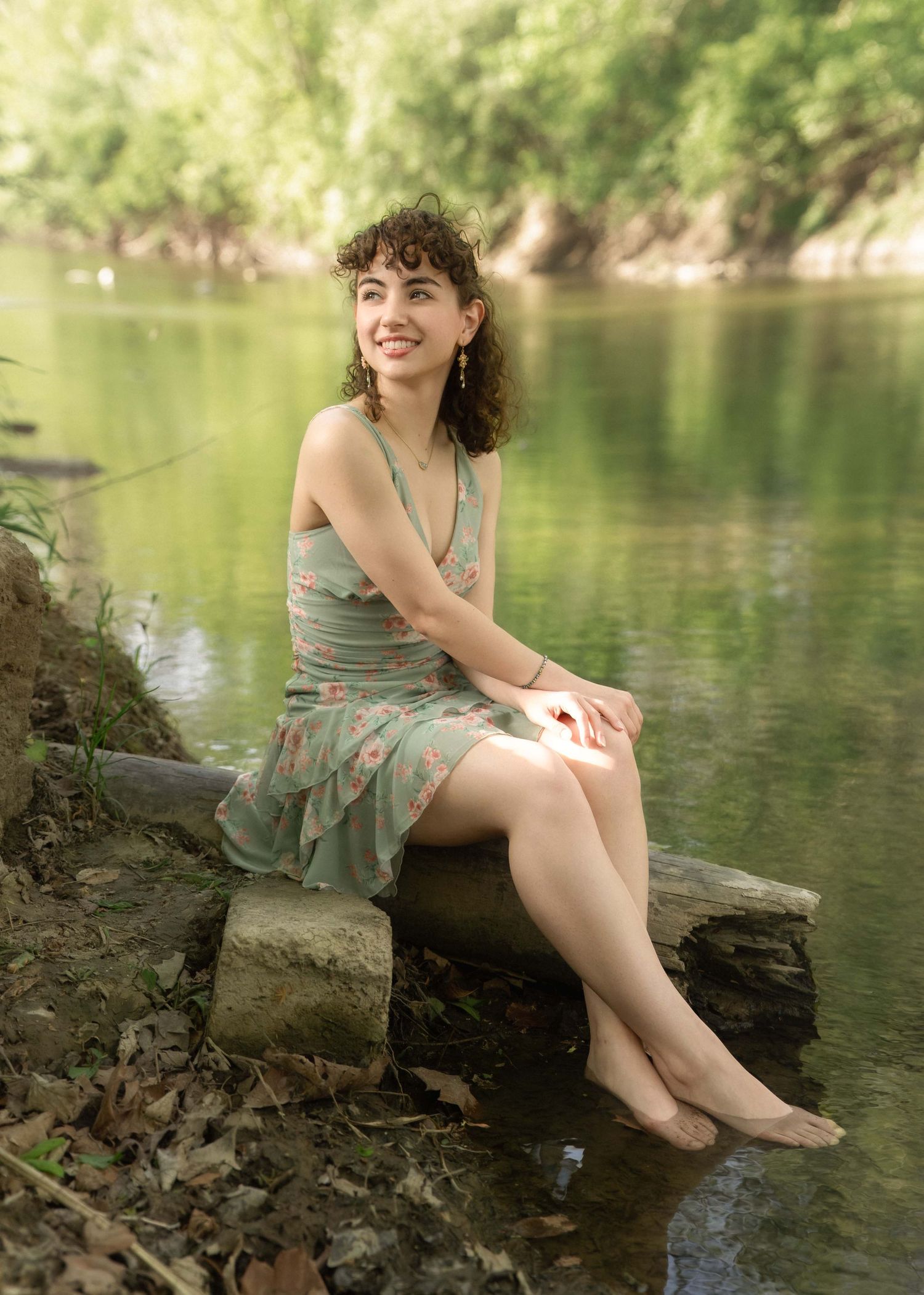 Young woman in green floral dress sitting barefoot on rocks by a serene tree-lined river, smiling outdoors.