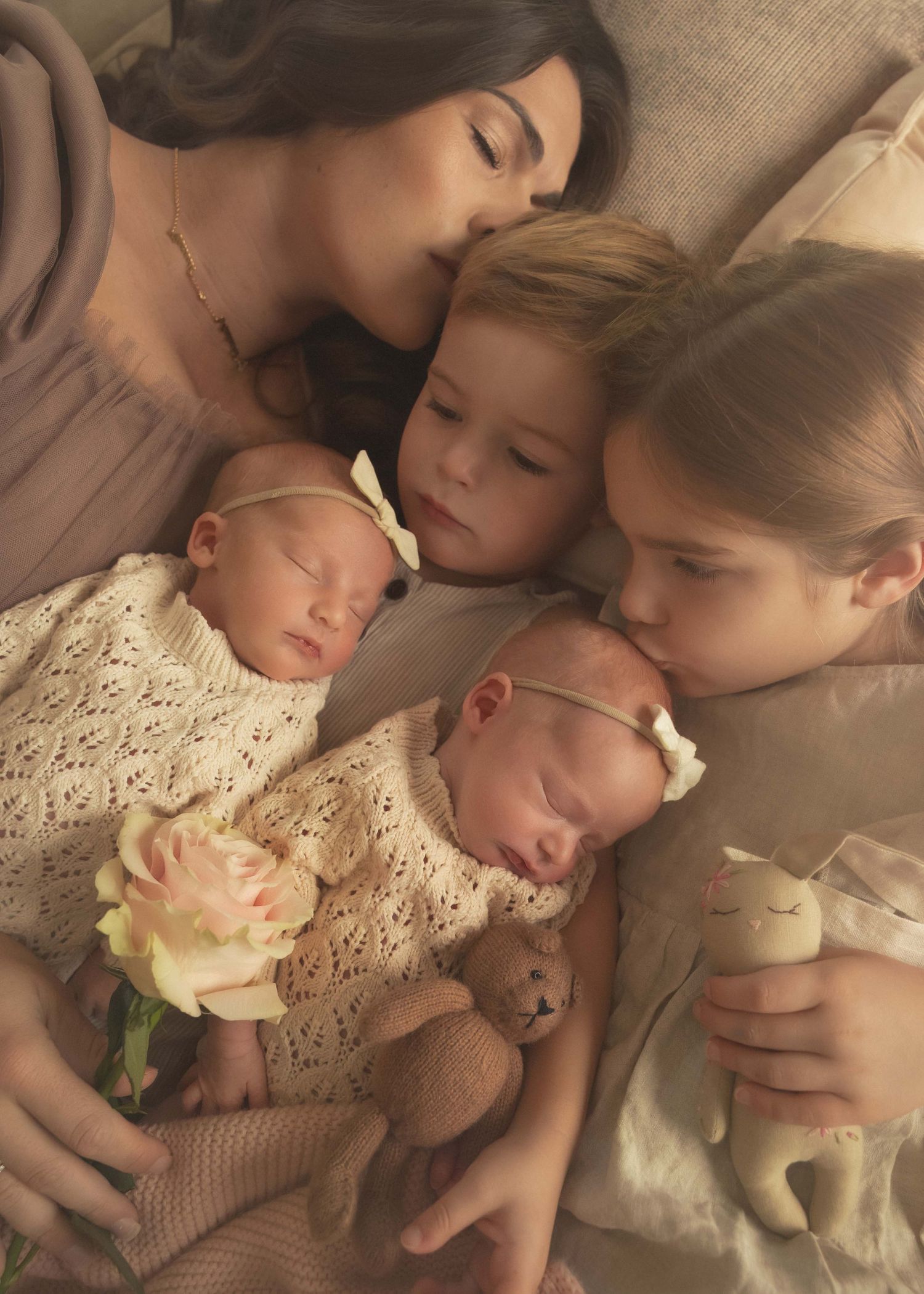 A mother tenderly kisses her toddler while two sleeping newborn twins in white knit outfits rest beside their siblings.