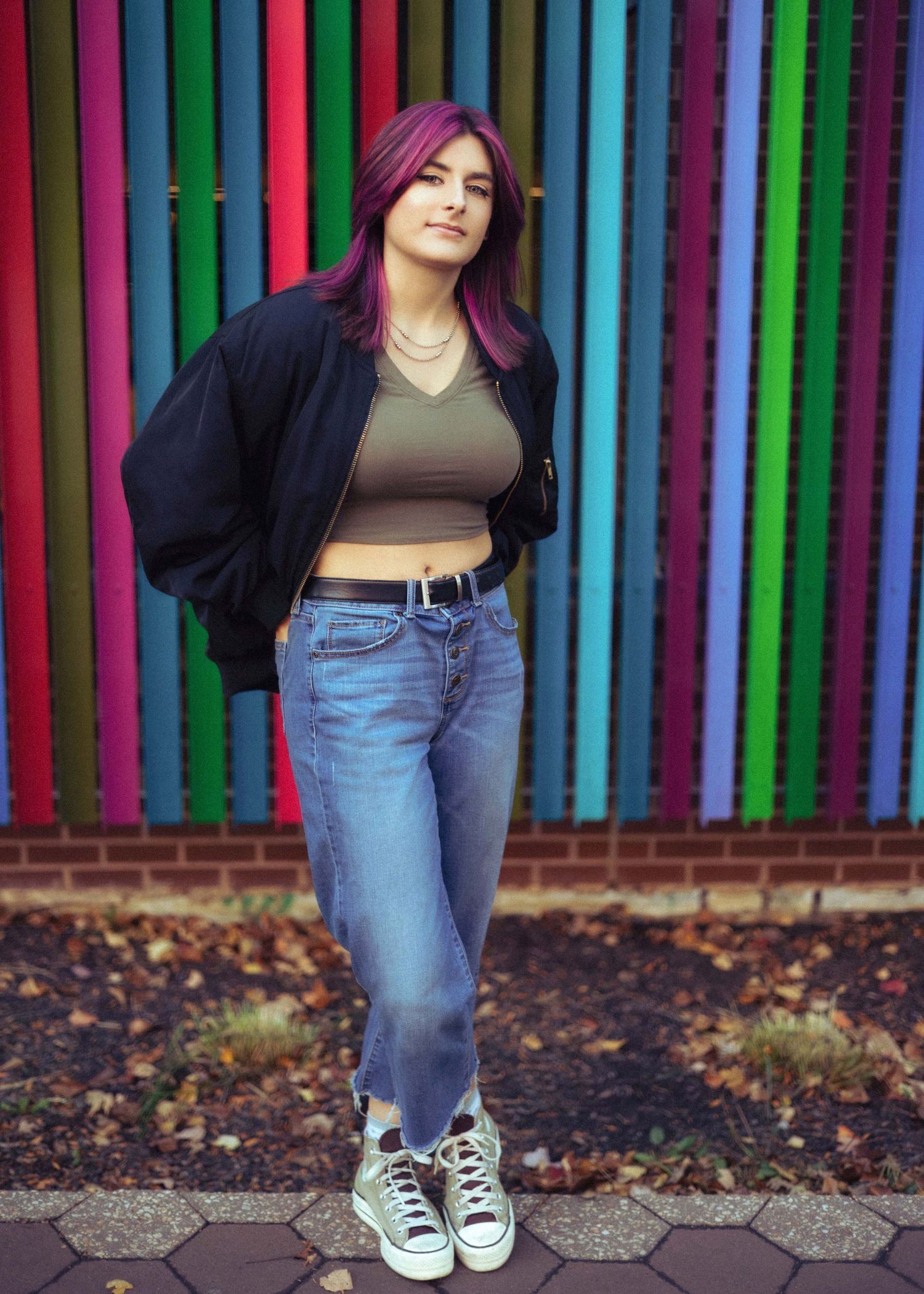 Young woman with purple hair in black bomber jacket, crop top, and jeans poses before colorful striped wall.